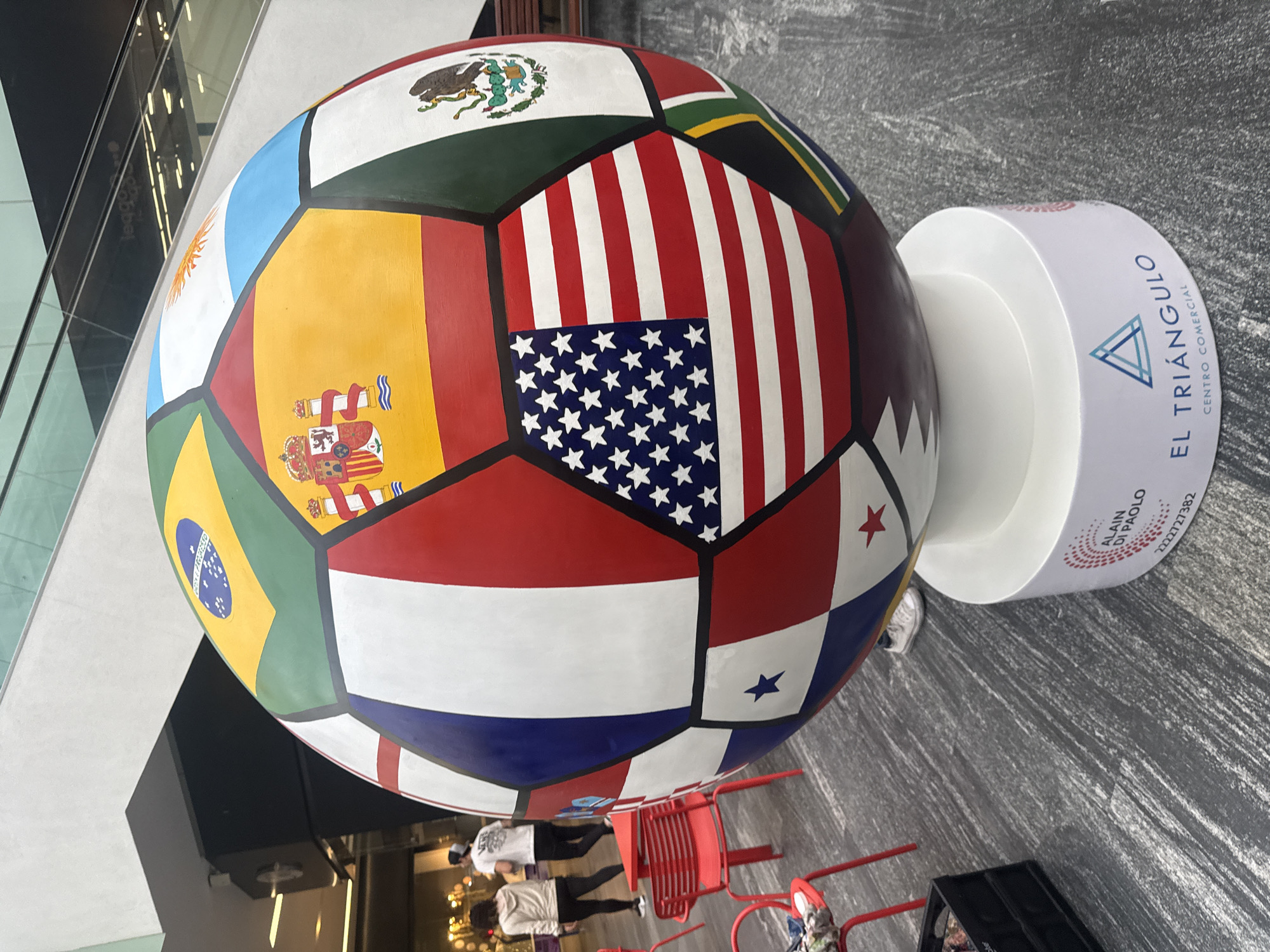 A large soccer ball sculpture sits on a white cylindrical base in the El Triángulo Centro Comercial in Puebla, Mexico. The ball is decorated with flags from various countries, including Mexico, Argentina, Spain, Brazil, USA, Panama, and South Africa. People are walking by red tables and chairs in the background. The area has a modern, commercial feel with gray-toned flooring and an open, well-lit space. A logo for "Alain Di Paolo" and the phone number 2222727382 are visible on the base of the sculpture.