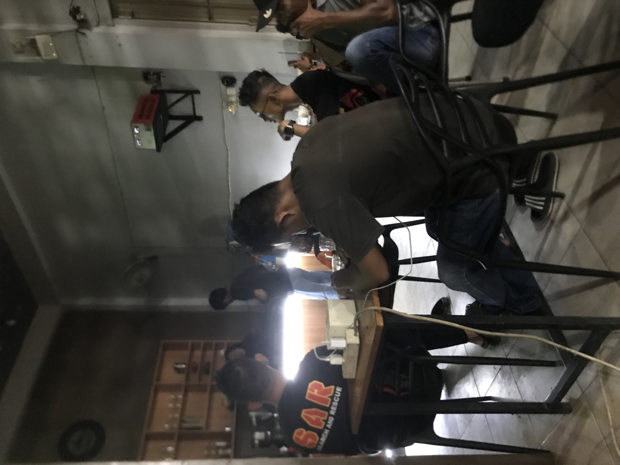 The image depicts an indoor scene, likely a cafe or restaurant in Lhokseumawe, Indonesia, with several people seated and engaged in activities involving electronic devices, potentially a collaborative working environment.
People:
There are multiple individuals visible, mostly young men. One is wearing a black shirt with "SAR" printed on it, another has glasses. A man in the background is wearing a hat.
Objects:
Several chairs are present, appearing to be metal-framed. A power strip and electrical cord are visible on the table. A bookshelf with various items can be seen in the background. A wall-mounted fan, lighting fixtures, and a fire safety system are also observed.
Actions/Interactions:
Individuals appear to be focused on their devices or on each other.
Environment:
The location is an enclosed space with tile flooring. The background suggests a public area with a bookshelf/display. The lighting is adequate.