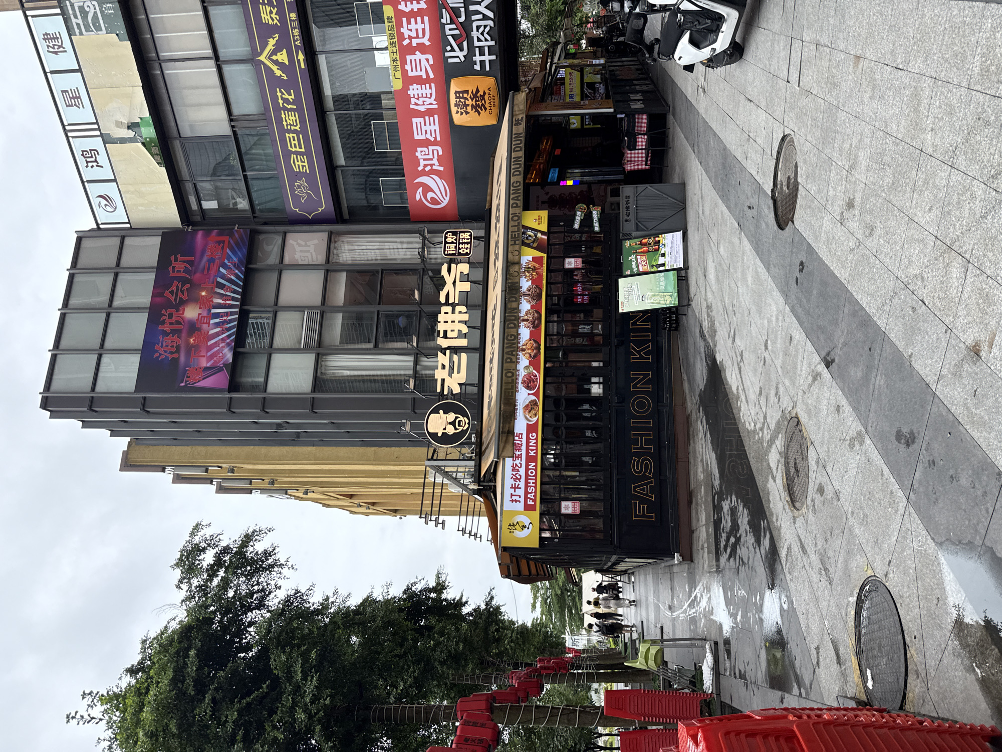 The image depicts a street scene in Haizhu District, China, on an overcast day. The foreground shows a paved sidewalk with manhole covers and puddles, indicating recent rain. To the left, stacks of red plastic chairs are visible. A building with a black facade prominently displays "FASHION KING" in large golden letters. Above this, a restaurant awning features various food images and the text "HELLO! PANG DUN DUN." The building itself is a multi-story structure with many windows. Red lanterns are hung along trees lining the sidewalk. Several people are walking away from the camera on the sidewalk, and a white scooter is parked on the right side of the street. Signs with Chinese characters and colorful graphics are displayed on the building's upper floors, indicating commercial establishments.