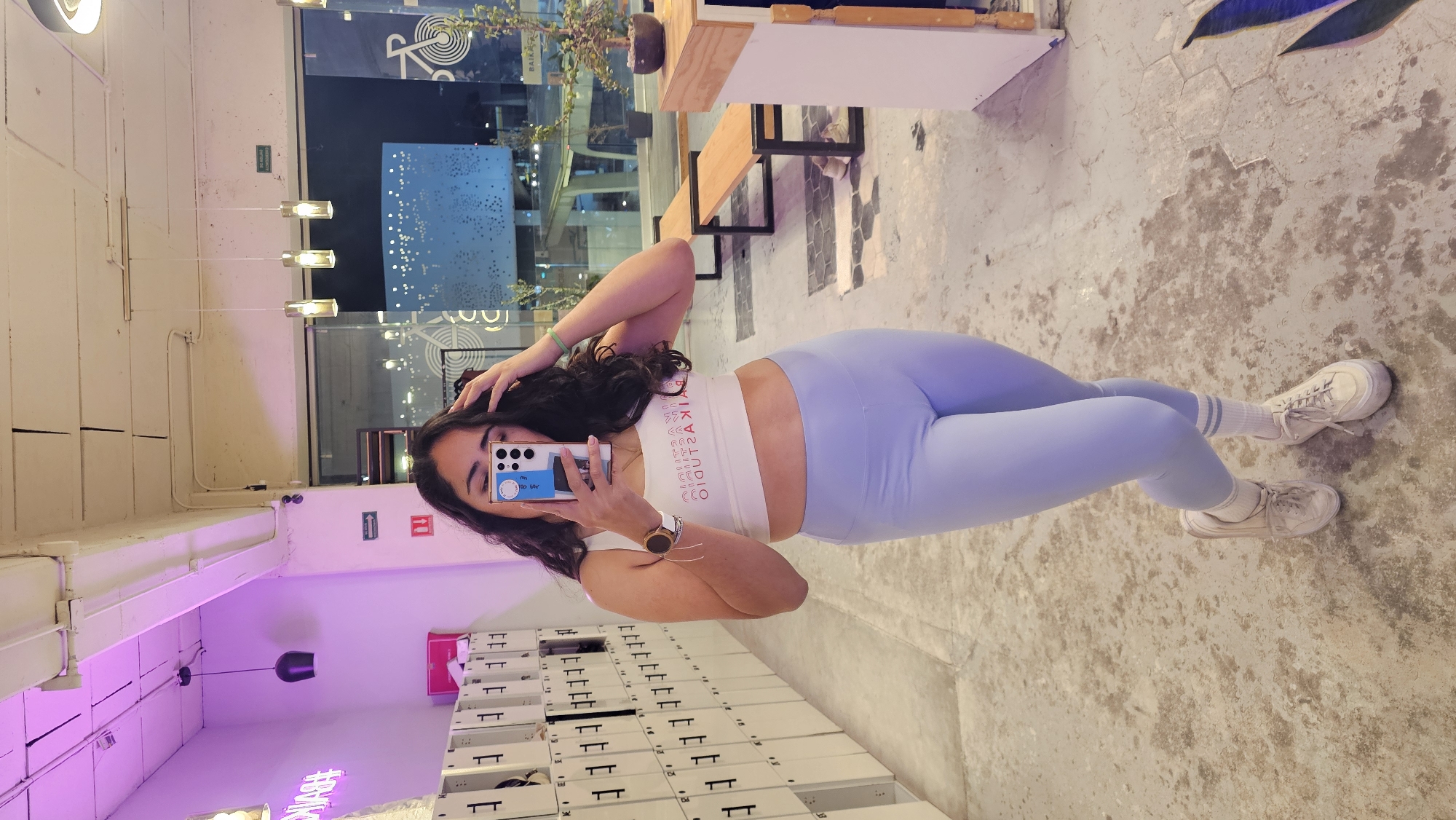 A female individual captures a mirror selfie inside a modern fitness studio at night. She wears a white crop top displaying the reversed text "BAIKA STUDIO," light blue leggings, white striped socks, and white athletic shoes. A smartwatch is on her left wrist and a green bracelet on her right. Her right hand holds a smartphone, partially obscuring her face, while her left hand touches her dark, curly hair.
The interior features a concrete floor, a long row of white lockers with black handles lining the left wall, and an exposed concrete ceiling with visible conduits and various light fixtures, including a pink neon sign and pendant lights. A red emergency exit sign and a fire extinguisher symbol are present on the wall, along with a green "SALIDA DE EMERGENCIA" sign above the windows.
Through large glass windows on the right, an outdoor night scene is visible. This includes two wooden benches with black frames, a potted plant on an indoor wooden counter, and a distant modern building with an illuminated, patterned facade. A large graphic of a bicycle and the "BAIKA" text are visible on the window glass, identifying the establishment as Baika Studio.
This setting is consistent with Baika Studio located in Lomas de Angelópolis, San Andrés Cholula, Puebla, Mexico.