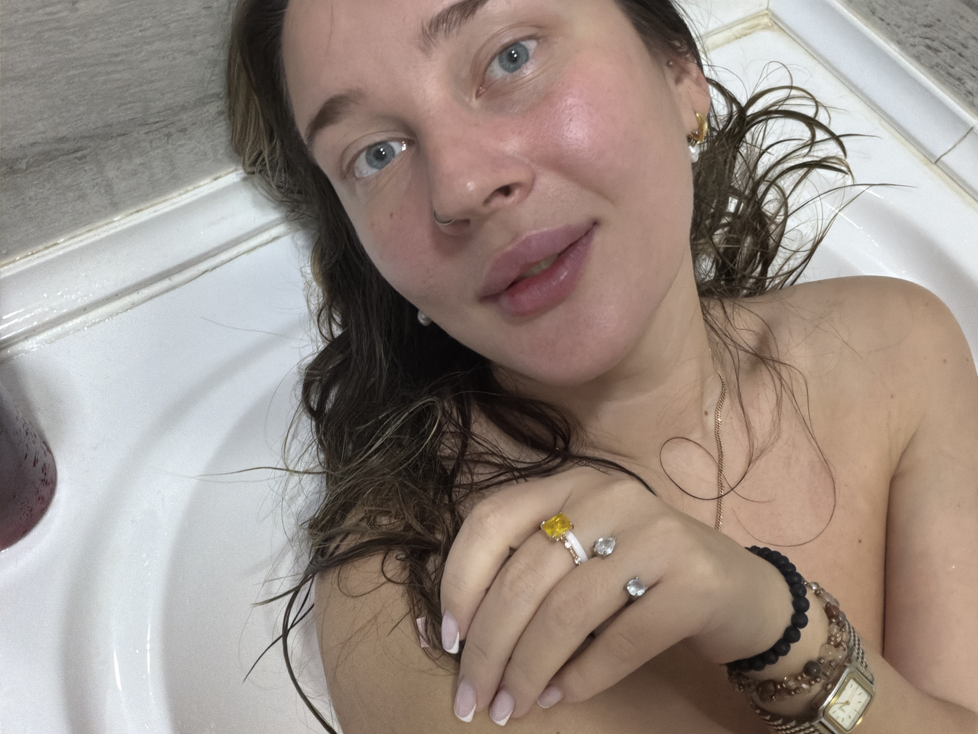 A woman is captured in a selfie while relaxing in a white bathtub, likely in a bathroom located in Chaikovsky, Russia. She is depicted from the chest up, with her wet, dark hair fanned out around her. Her striking blue eyes look directly at the camera, and she wears a subtle, relaxed smile. A silver nose ring adorns her nostril, and a gold hoop earring with a pearl drop is visible in her right ear. A thin gold chain necklace rests on her bare chest.
Her right hand is gently placed near her chest, displaying several pieces of jewelry: a prominent ring with a large rectangular yellow stone on her ring finger, and two rings featuring clear stones (one round, one pear-shaped) on her middle finger. Her fingernails are manicured with French tips. On her right wrist, she wears multiple beaded bracelets, including black and earth-toned beads, alongside a vintage-style watch with a square, light-colored face. A dark red bottle is partially visible to the left in the tub. The background shows light grey wall tiles and the edge of the white bathtub, which appears to have some minor discoloration along the rim. The overall scene conveys a casual and private moment of relaxation.