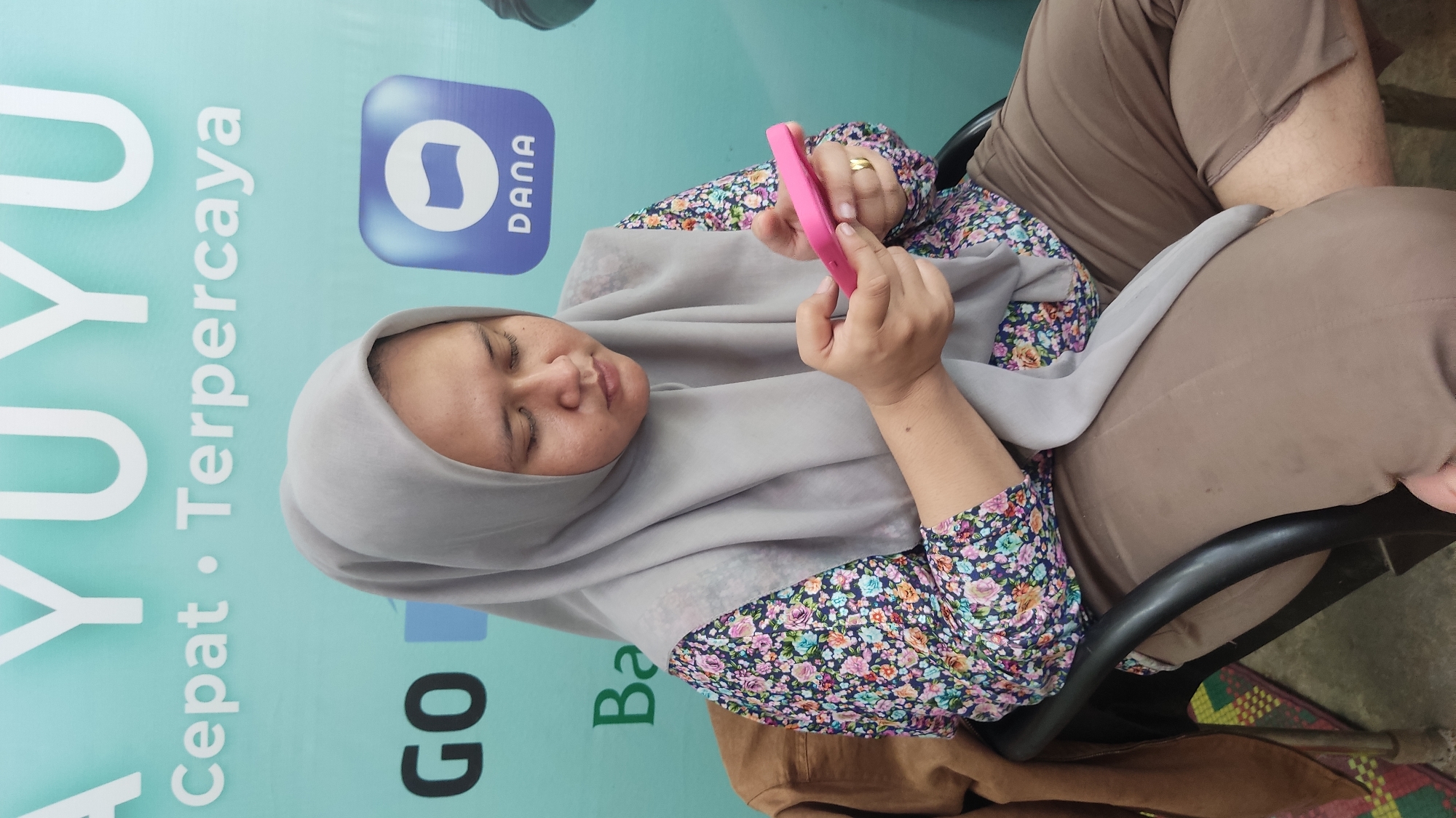The image, rotated 90 degrees clockwise, captures a woman seated in what appears to be an indoor or shaded outdoor setting. She is wearing a light grey hijab, a dark blue floral long-sleeved top with pink, white, and green patterns, and light brown pants. She sits in a chair, with a black armrest visible.
The woman is focused on a bright pink smartphone, which she holds with both hands. Her gaze is directed downwards at the phone's screen, and her eyes appear partially closed, suggesting intense concentration or perhaps weariness. A gold-colored ring is visible on her right ring finger.
The background is dominated by a light blue wall or banner. Prominently displayed are large white letters forming the word "UYU," followed by "Cepat • Terpercaya" (Fast • Trustworthy in Indonesian). To the right, a blue square logo with the word "DANA" in white is visible. Below "Terpercaya," parts of words like "GO" and "Ba" (likely from "Bayar" or similar) can be seen, indicating an association with a payment or digital wallet service, consistent with the "DANA" logo. A patterned mat or rug is partially visible on the ground at the bottom right. The overall scene suggests the woman is using a mobile application, possibly for transactions or communication, within a business or public space in Lhokseumawe, Indonesia.