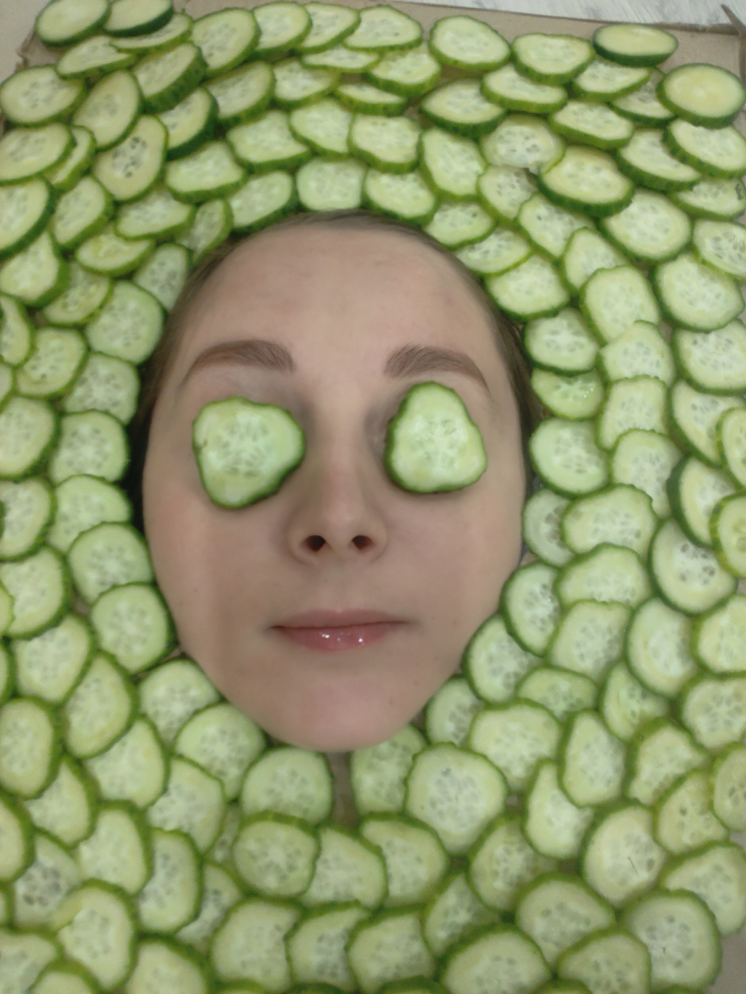 A woman's face is visible, surrounded by slices of cucumber. Two slices of cucumber are placed over her eyes. Her skin tone is fair. The setting appears to be indoors.