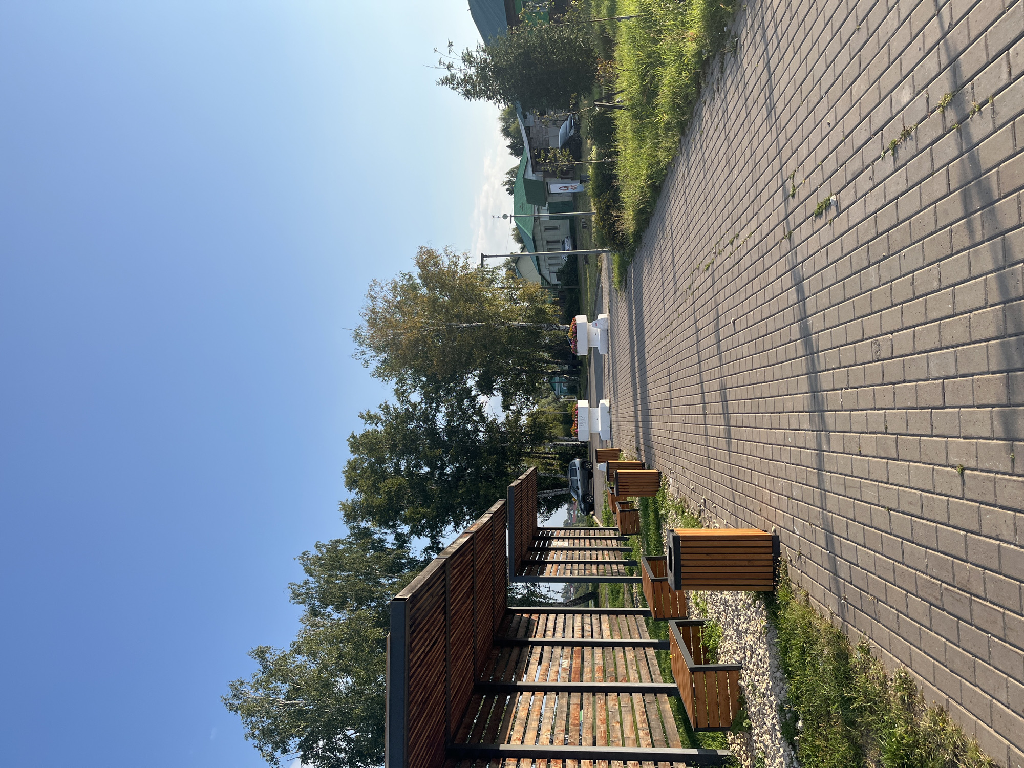 An outdoor paved walkway in Ufa, Russia, is lined with wooden benches and trash receptacles under a covered wooden structure. Tall green trees and houses with green roofs are visible in the background. The sky is clear and blue.