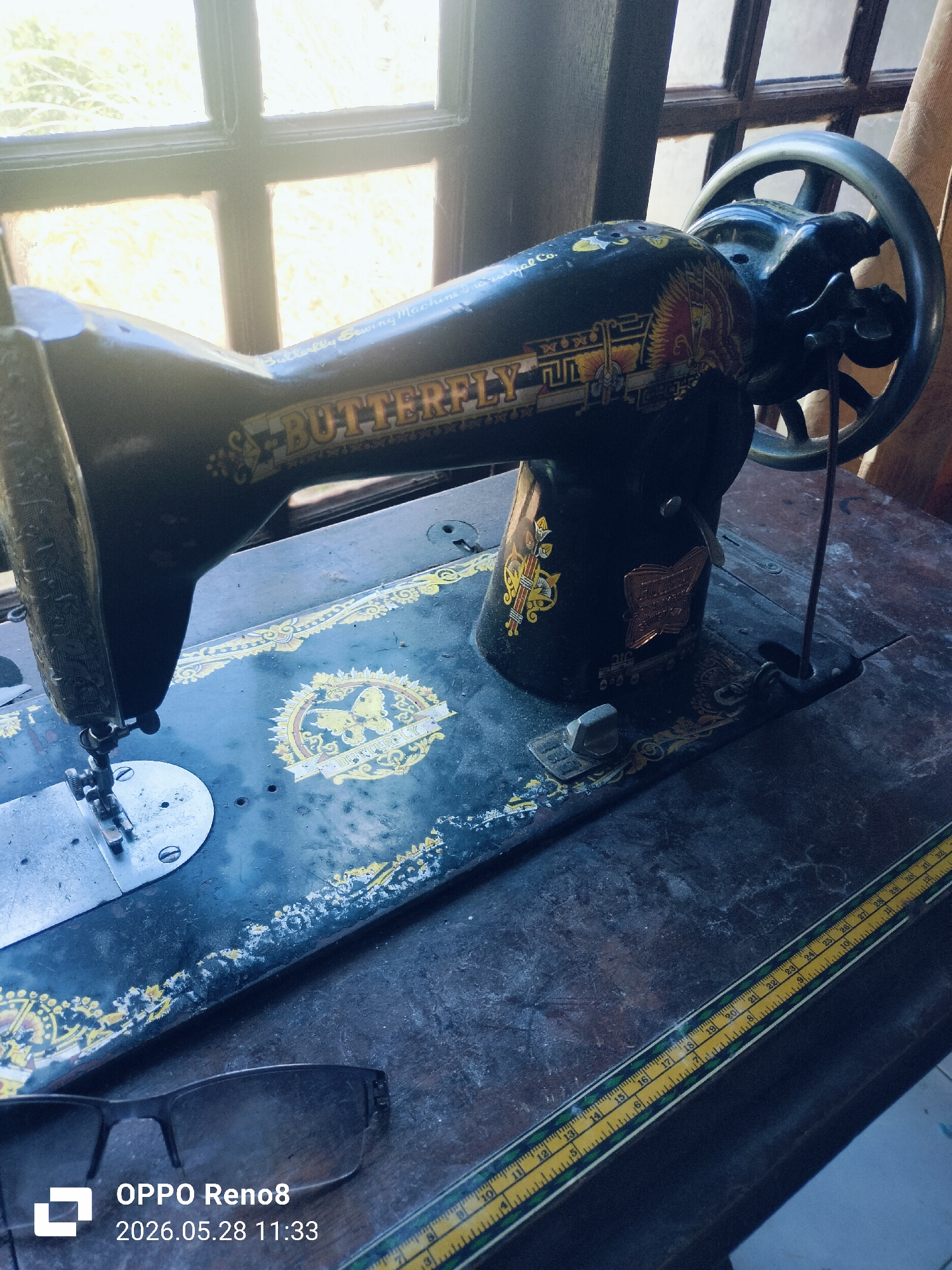 The image features an antique "Butterfly" brand sewing machine, positioned on a dark wooden surface near a window. The sewing machine is predominantly black with gold decorative accents and the brand name prominently displayed. To the lower left of the sewing machine are a pair of glasses. Along the lower right, a yellow and green ruler stretches across the table surface. The window behind the machine allows diffused light, suggesting an interior setting. The lower left corner of the image contains a digital watermark with the text "OPPO Reno8" and the date "2026.05.28 11:33." There are no people visible in the image. The location is not specified in the image or the prompt.