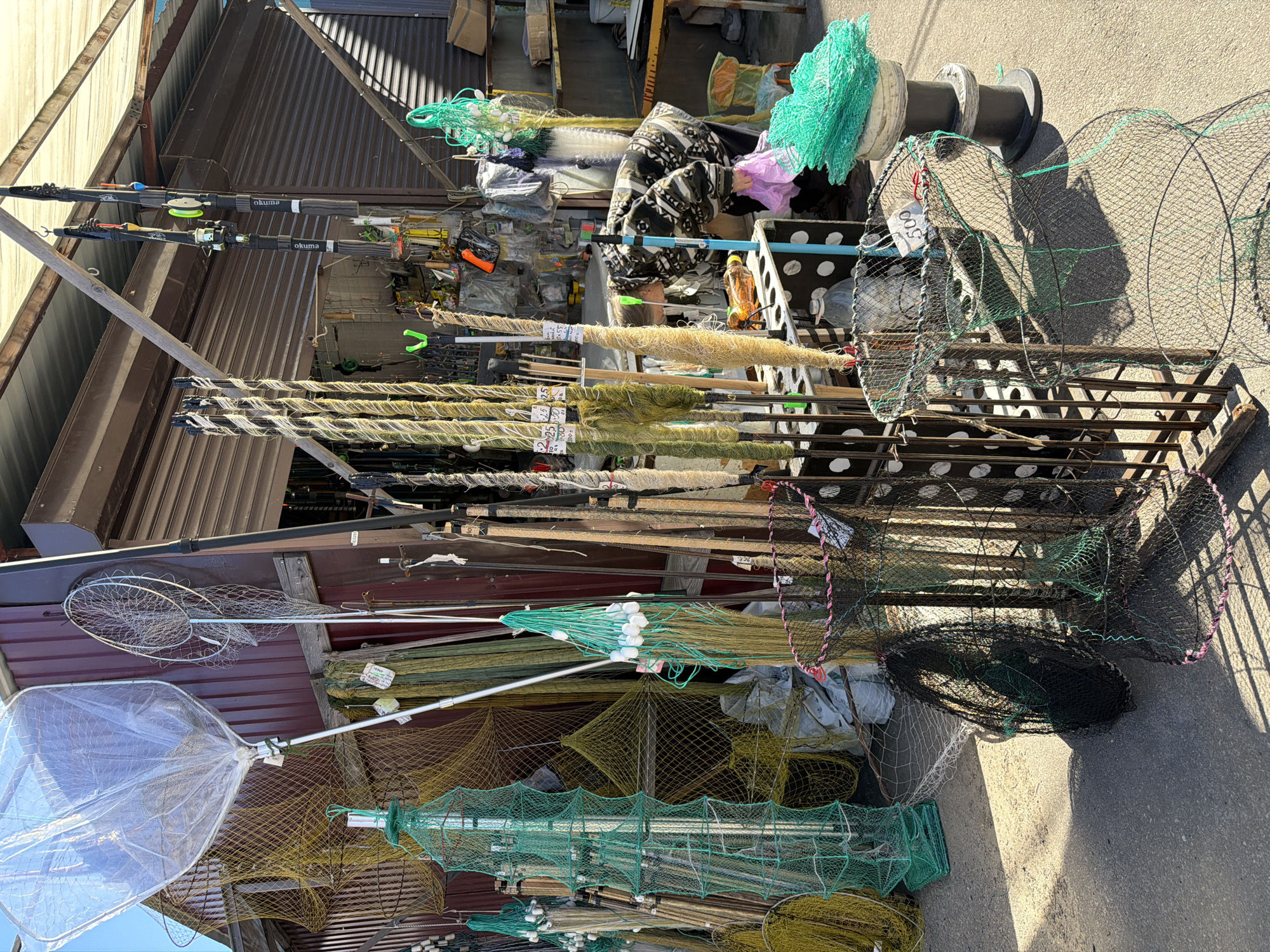 The image depicts an outdoor stall selling fishing equipment in Nizhny Novgorod, Russia. The stall features a variety of fishing nets, including large hoop nets, conical traps, and smaller nets with floats. Several fishing rods, some with reels attached, are also displayed. A woman wearing a patterned jacket is browsing the merchandise. The background shows a building with a corrugated metal facade and an awning, suggesting a market or a roadside stand. The ground is paved, and sunlight casts shadows, indicating it is daytime.