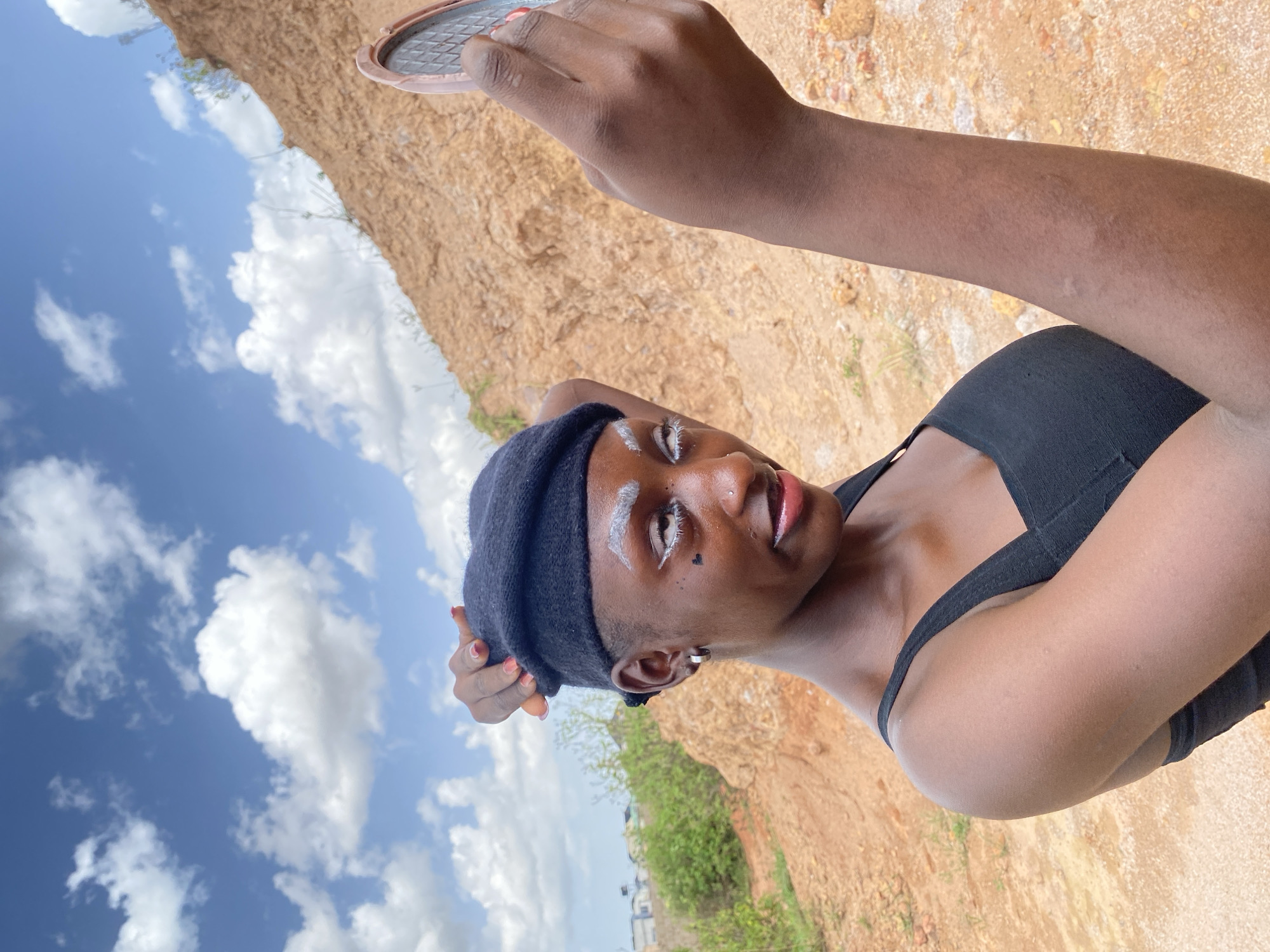 This media file shows a person in a black tank top and a black beanie. The person has white makeup on their eyebrows and a small heart-shaped marking on their cheek. They are holding a compact mirror in their right hand, which is raised towards the camera. The background consists of a sandy, rocky landscape with some greenery and a bright blue sky with scattered white clouds. The image appears to be taken outdoors, likely during the daytime, given the lighting and the sky. The overall mood seems casual and playful. There is no visible text in the image. The location context provided is Bar Jirgi Summa, Nigeria.