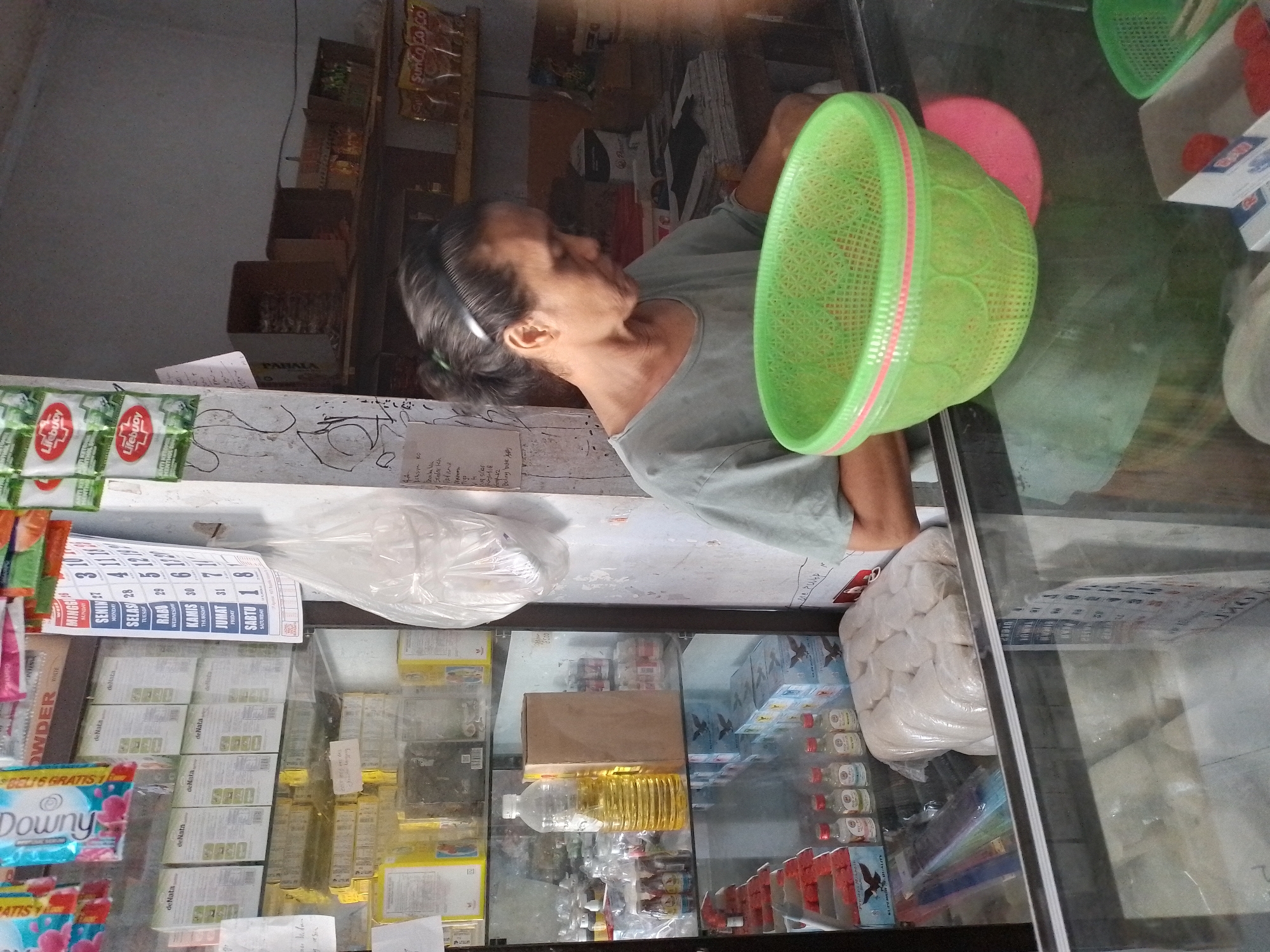 A middle-aged Indonesian woman with dark hair pulled back in a ponytail is standing behind a glass display counter in what appears to be a small convenience store or warung in Ngancar, Indonesia. She is wearing a light gray t-shirt and is looking down and to her right, possibly at an item on the counter or preparing something. In front of her on the counter is a bright green plastic basket with a pink trim and a pink circular object underneath it, also on the counter. The background shows shelves stocked with various goods, including bottles of oil, boxes, and packaged items. A calendar is visible on a pole to the woman's left. The overall lighting suggests an indoor setting, possibly during the daytime.