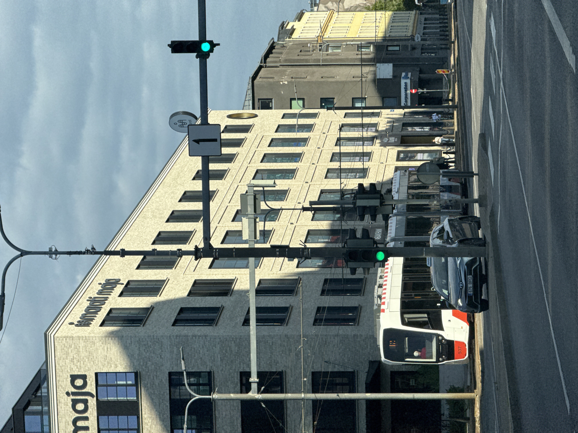 The image depicts a street scene in Tallinn, Estonia, likely in the city center given the presence of modern architecture and public transportation. In the foreground, a dark-colored sedan with a license plate "676 BRD" is parked or stopped at an intersection. Behind the car, a modern tram, number 537, with red and white accents, is visible. The tram has its doors closed, and a person appears to be in the driver's seat.
Dominating the background is a multi-story building with a light beige brick facade, bearing signage that reads "maja" and "ismaroti maja". The building features numerous windows, some with dark shutters. Adjacent to this building, a darker, taller structure stands, followed by a row of buildings with yellow and lighter colored facades.
The street is paved, with visible lane markings and pedestrian crossings. A traffic light at the intersection is illuminated green. A street lamp pole stands to the left of the tram, and another, more complex pole with traffic lights and signage, is to the right of the building. There are several pedestrians visible on the sidewalk in front of the buildings. The sky above is cloudy, with patches of blue sky visible. The overall impression is a contemporary urban environment.