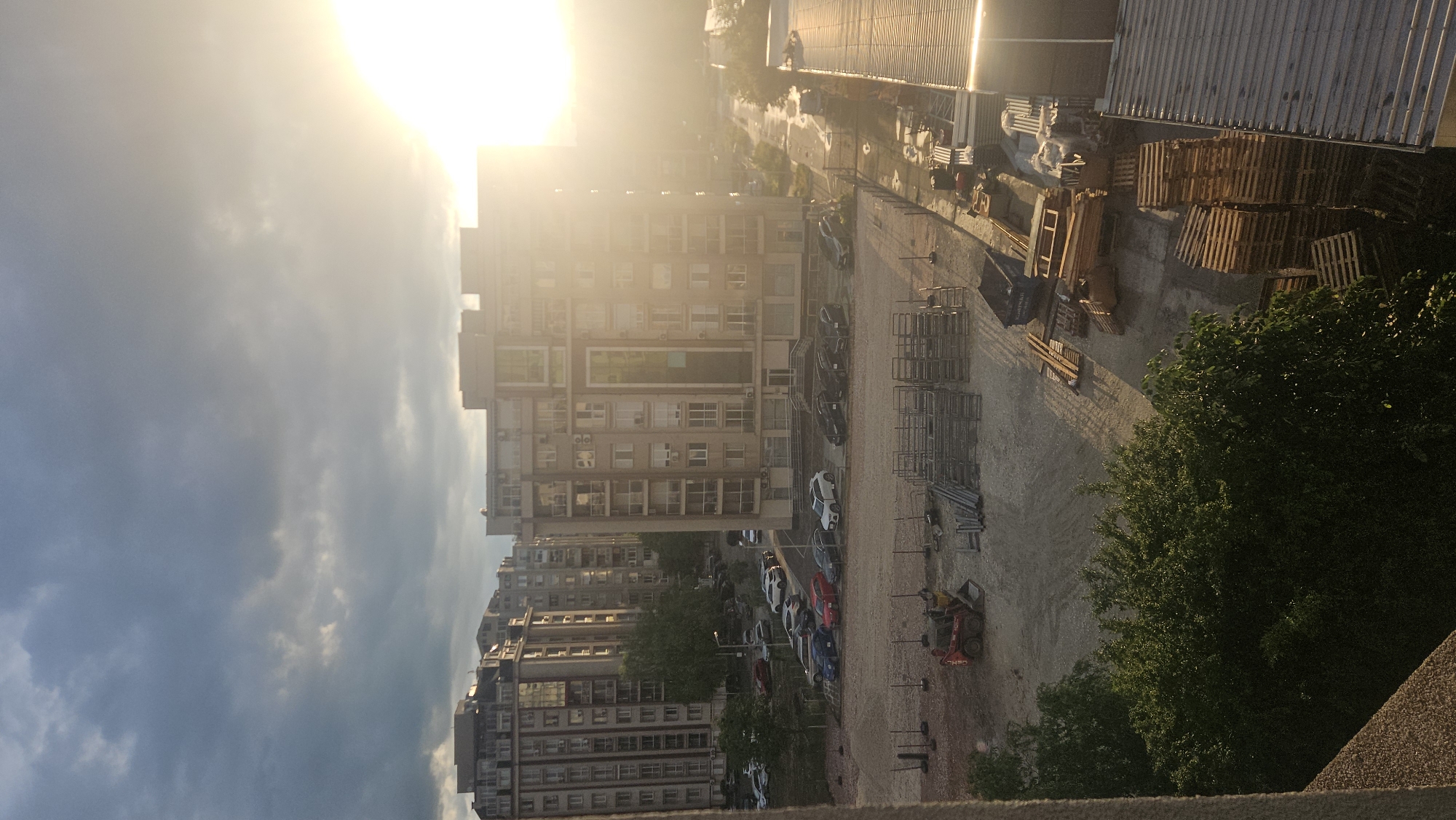 The image depicts an urban scene in Plovdiv, Bulgaria, viewed from an elevated position. Multiple buildings, varying in height and architecture, are visible. A parking lot with several cars is located between the buildings. Construction materials, including wooden pallets and metal frames, are scattered in an open area adjacent to the parking lot and some trees are present. The upper portion of the image is dominated by bright sunlight and sky with clouds.