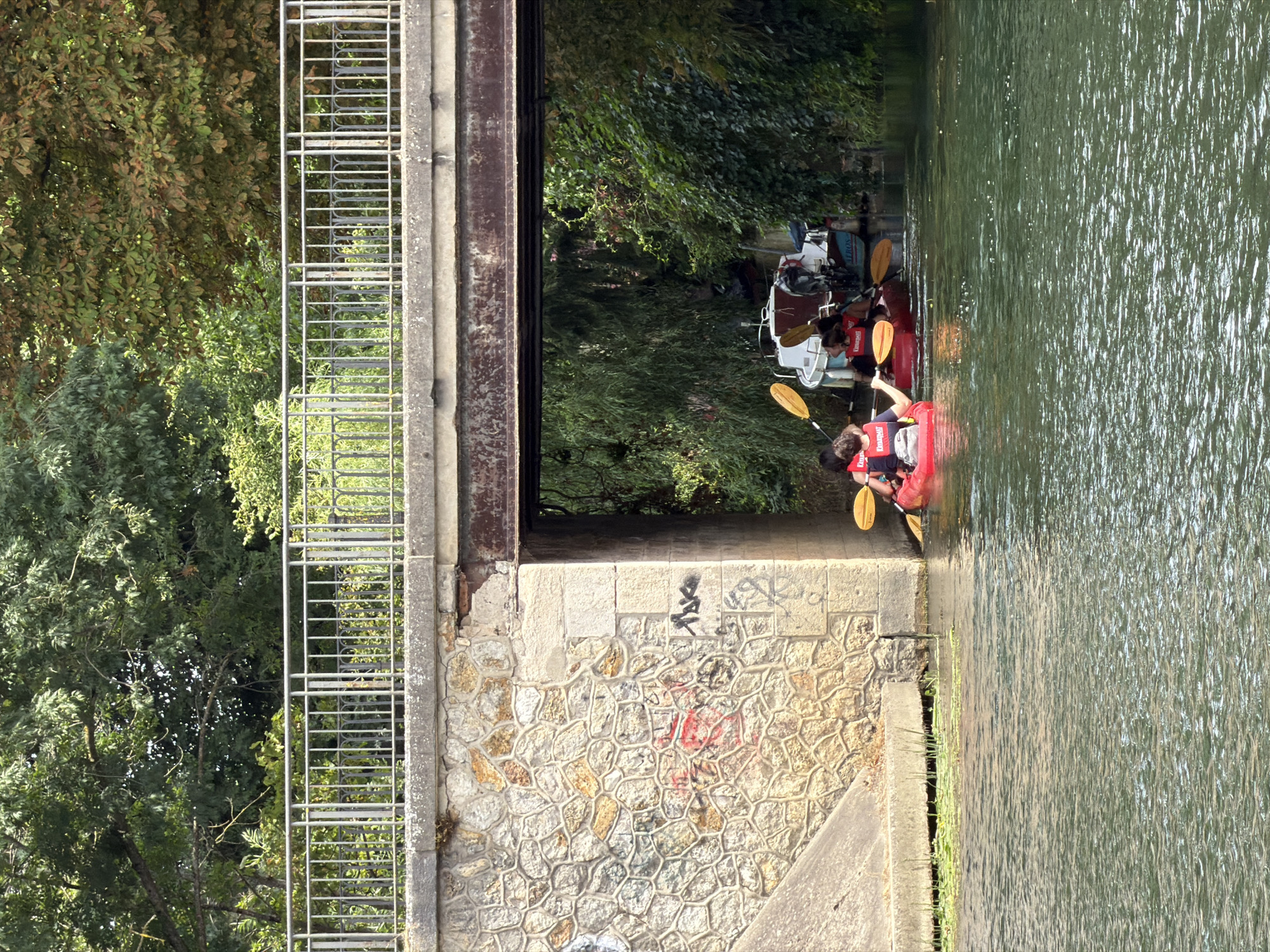 The image depicts a bright, sunny day on the Marne River in Joinville-le-Pont, France, beneath a stone and concrete bridge. Two red kayaks are visible in the foreground, each carrying two individuals wearing life vests. The kayakers are paddling, suggesting an activity or sport. Behind the kayaks, two moored boats are partially visible, adding to the scene's context as a waterway. Lush green foliage from trees and bushes borders the riverbanks and overhangs the bridge, creating a natural, vibrant environment. The bridge itself features a metal railing and appears to have some graffiti on its stone supports. The water is a murky green, reflecting the surrounding greenery.