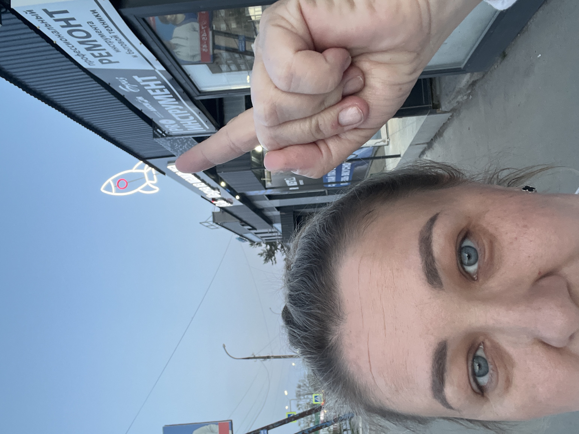 A selfie taken in Chaikovsky, Russia shows a woman pointing at a storefront with a rocket-shaped light. The storefront sign reads "ПРО ИНСТРУМЕНТ" and "РЕМОНТ." The woman is in the foreground, with the storefront and street behind her. The sky is light blue.