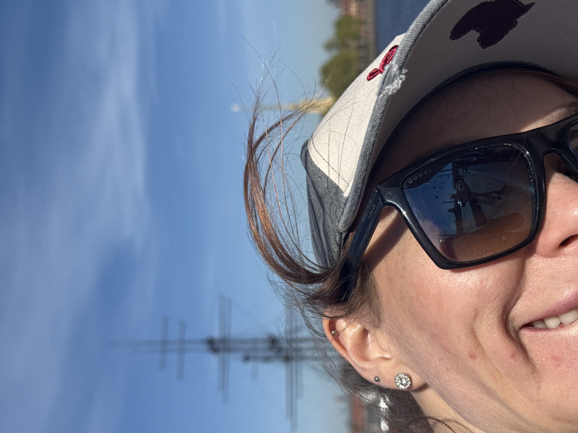 The image shows a partial view of a woman wearing a baseball cap and Gucci sunglasses. The sunglasses reflect an image of a sailing ship, indicating a possible location near a harbor or waterway. In the background, the masts and rigging of a ship are visible against a blue sky. Based on these elements and the location provided, this photo likely depicts the woman near the Neva River in Saint Petersburg, Russia, possibly during a visit to a maritime-related landmark or event.