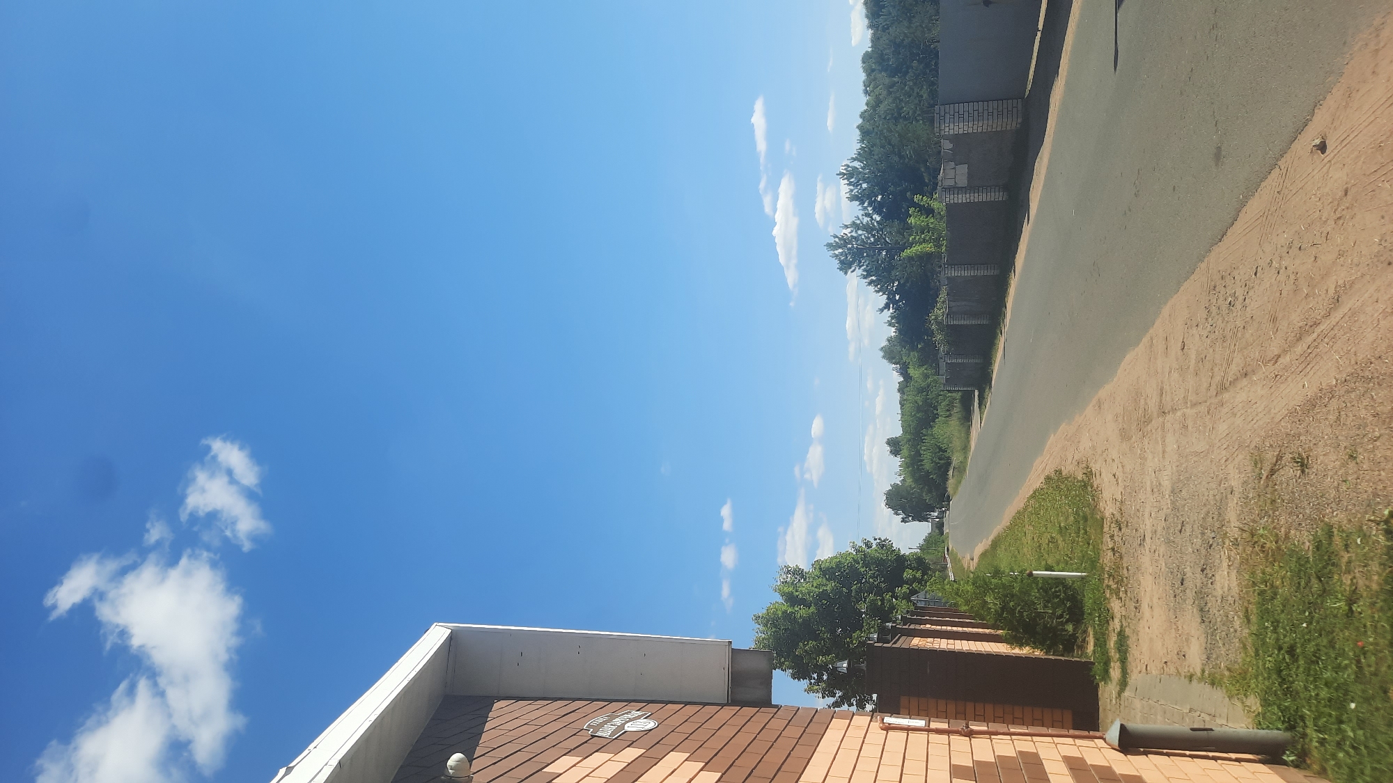 The image shows a rural road in Shklov, Belarus, on a clear day. The road is paved and runs through a wooded area with fences on either side. There is a brick house on the left side of the road, with a white roof overhang. The sky is blue with scattered white clouds. The environment appears peaceful and quiet, with no people visible in the frame.