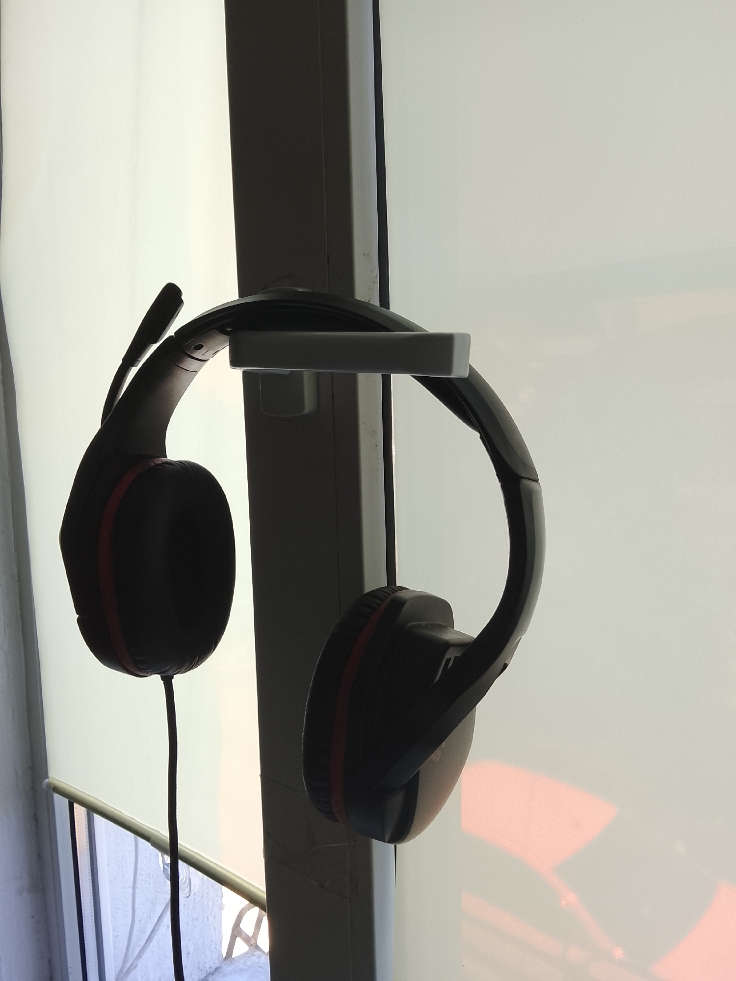 A pair of black gaming headphones with red accents hangs from a white mount attached to a window frame. The headphones have a microphone extending from the left earcup. The background is a sheer white roller blind illuminated by soft, diffused light, suggesting it is daytime, possibly overcast or early morning/late afternoon. The blind is positioned in front of a window, with glimpses of a window frame and possibly an exterior view, including a balcony railing, visible through the lower portion of the blind. The lighting creates a silhouette effect on the headphones, obscuring finer details and casting soft shadows. The overall scene is simple, focusing on the hanging headphones as the primary subject. No text is visible in the image. The setting appears to be an indoor environment, likely a room with a window.