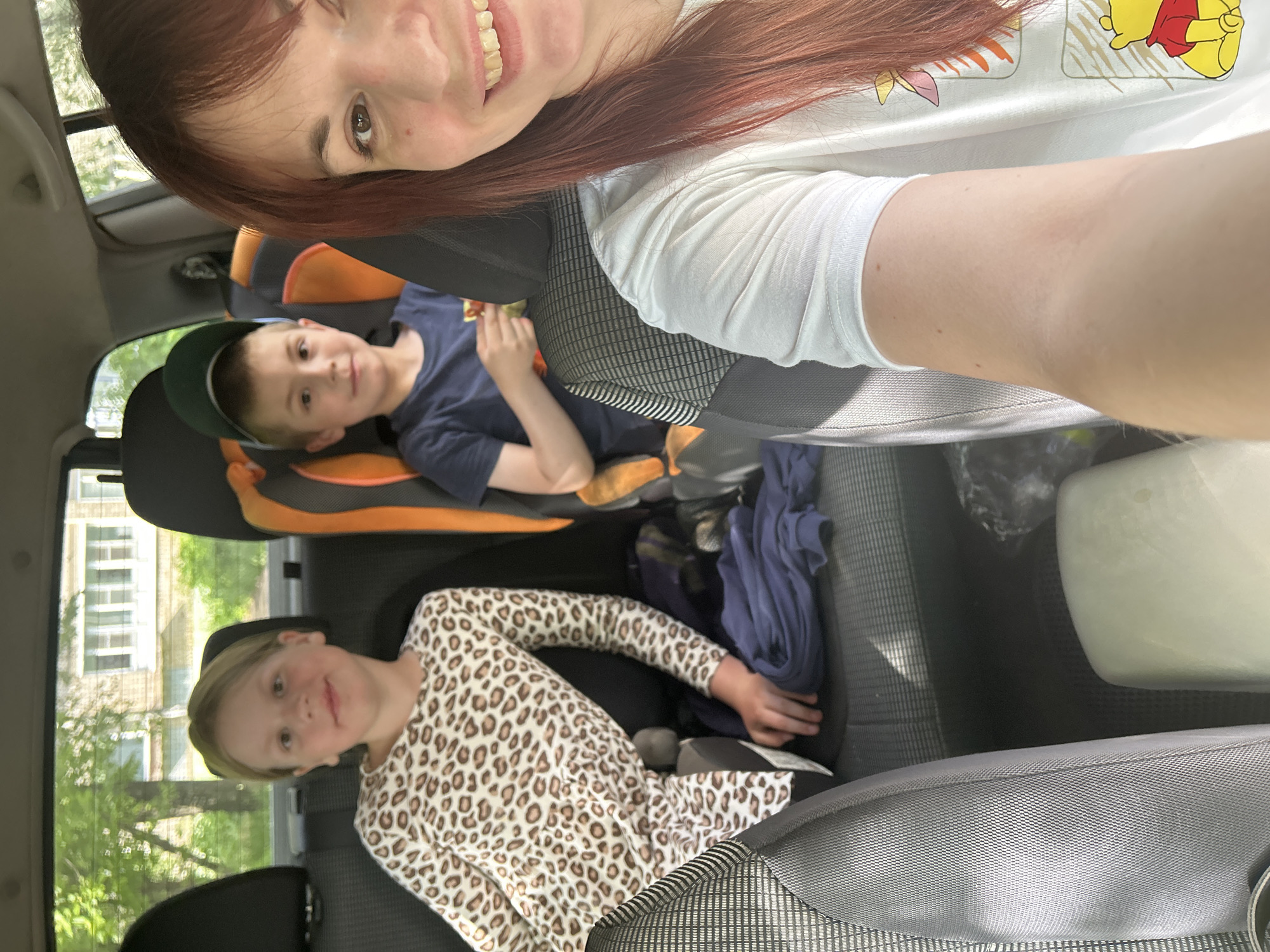 A woman with long red hair is taking a selfie in the front seat of a car. Behind her, two children are visible in the back seat. One child is a boy in a car seat wearing a baseball cap and a blue shirt. The other child is a girl in a leopard-print dress. The car is driving past a building with windows, and trees can be seen outside. The location may be in the city of Chaikovsky, Russia.