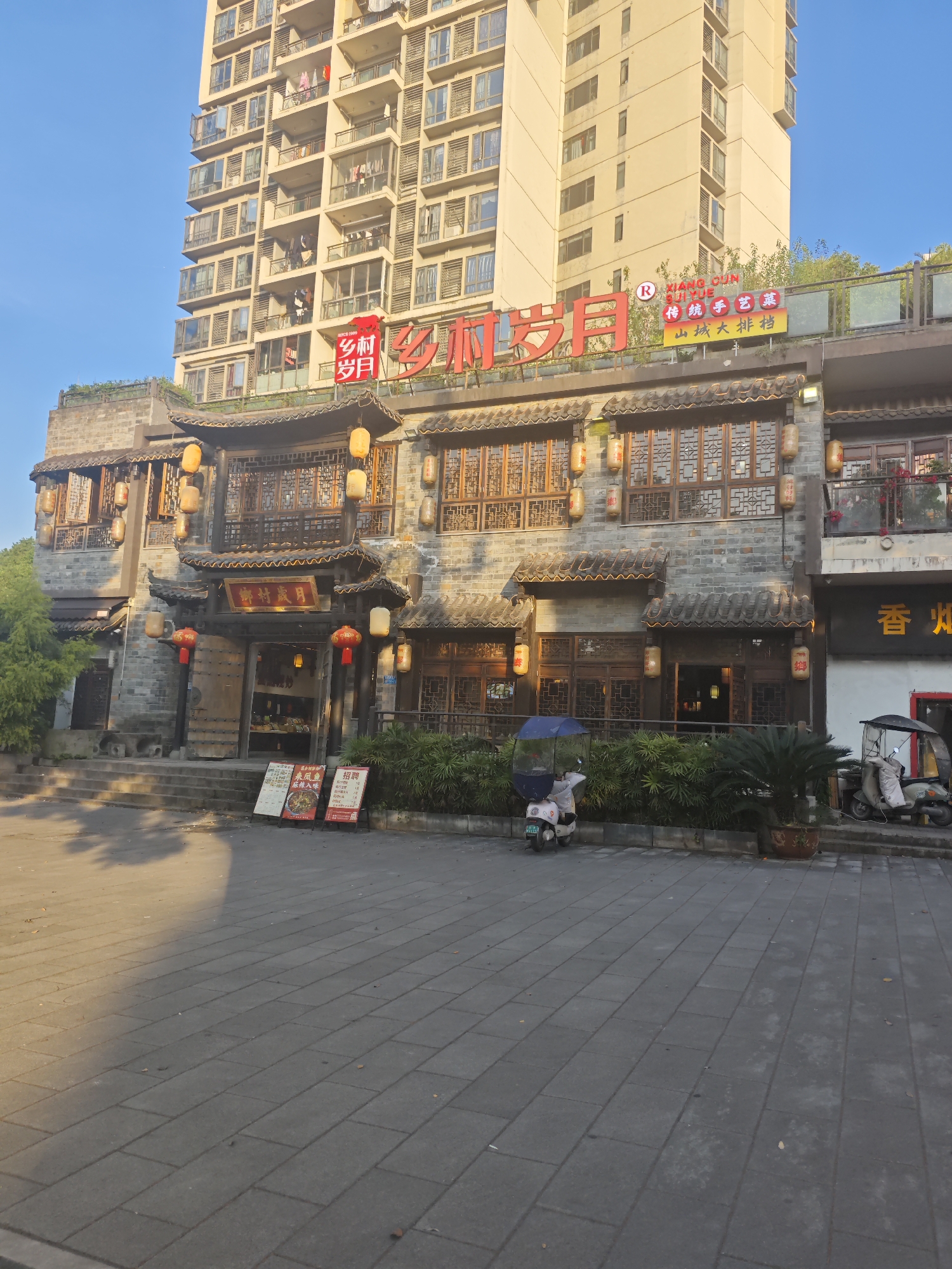The image shows a restaurant called "Xiang Cun Sui Yue" in Chongqing High-tech Zone, China. The restaurant is designed with traditional Chinese architecture, featuring gray brick walls, wooden windows, and red lanterns. Above the restaurant's entrance, a large red sign displays the restaurant's name in Chinese characters. A modern high-rise building rises in the background, contrasting with the traditional design of the restaurant. Two scooters parked in front of the building. A few trees and plants add greenery to the scene. The foreground consists of a tiled pedestrian walkway, with part of the area illuminated by sunlight, creating shadows.