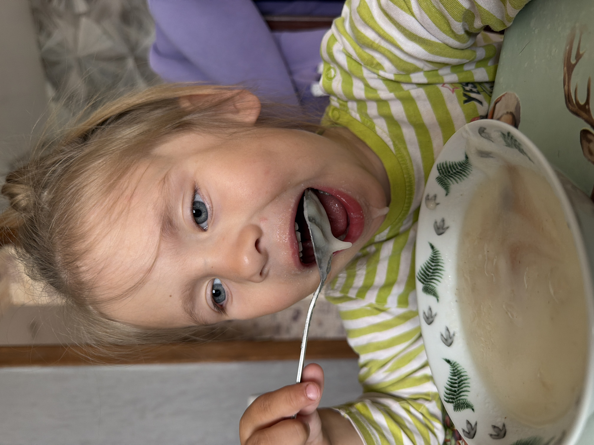 A close-up, eye-level shot shows a young blonde girl with blue eyes eating from a bowl of soup. She is wearing a green and white striped long-sleeved shirt. Her mouth is open wide as she takes a spoonful of soup, with some soup dripping from her chin. The bowl has a decorative pattern of green ferns and gray footprints around the rim. The background is slightly blurred, showing a neutral-colored wall and a hint of purple fabric. The girl's hair is pulled up in a messy bun. The scene appears to be indoors, likely in a home environment, and is set in the city of Chaikovsky, Russia.