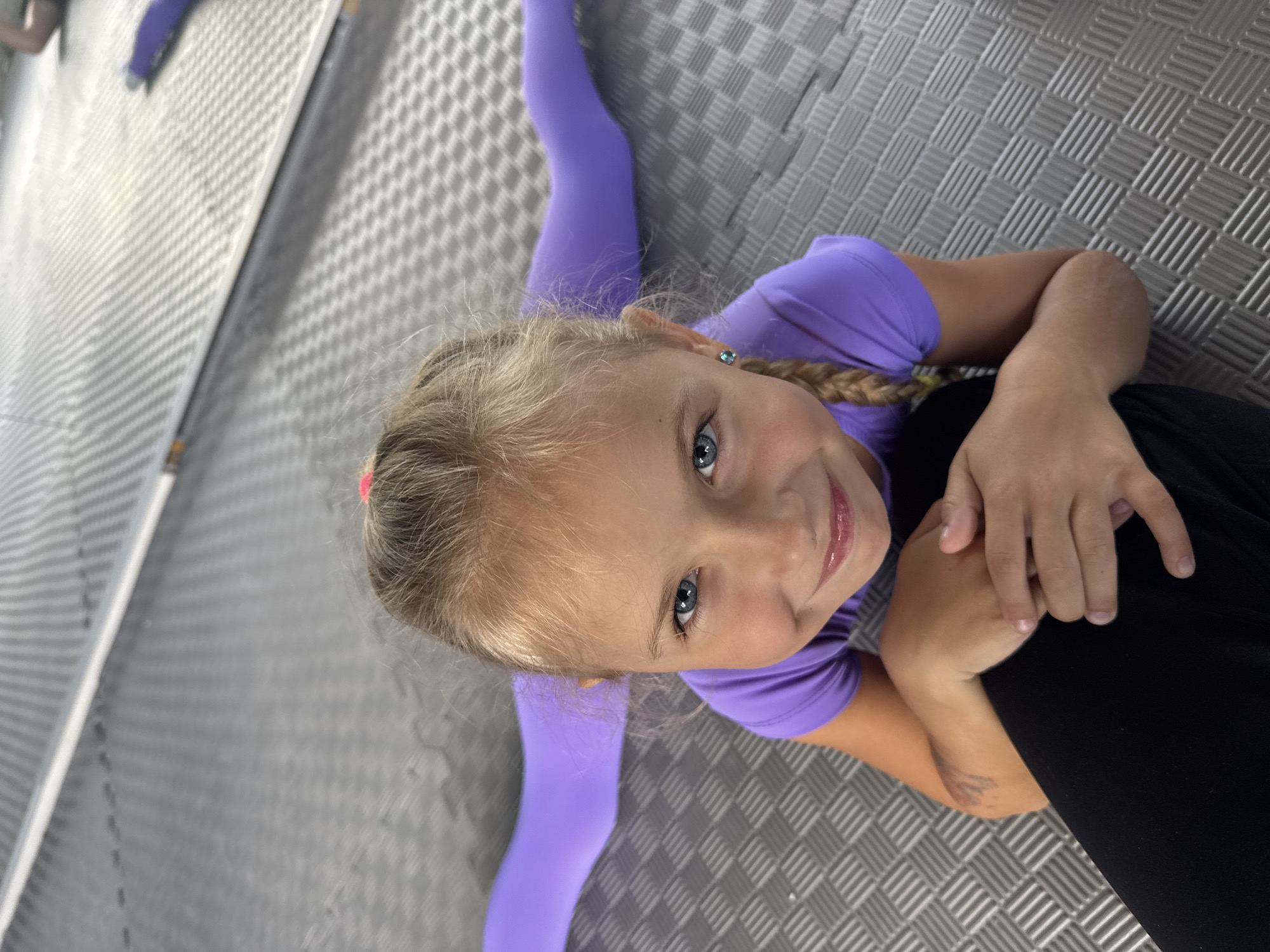 A young girl with blonde hair in a braid and blue eyes is in a split on a grey, textured floor. She is wearing a purple leotard and purple leggings. The girl is looking up at the camera with a smile. Her left hand is resting on her left knee, and her right hand is on her right thigh. In the background, there is a reflective surface, possibly a mirror or a wall. The lighting suggests an indoor setting, possibly a dance studio. The girl appears happy and confident. There is no visible text in the image.