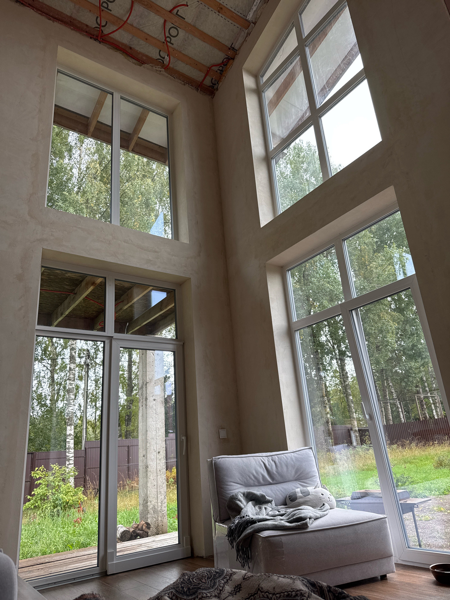 The image depicts the interior of a spacious room with large windows overlooking a wooded outdoor area. The room appears to be under construction or recently renovated, with exposed ceiling joists and insulation visible in some areas. The walls are textured and light-colored, and the floor is made of wood.
In the foreground, a gray upholstered chair with a knitted throw blanket and a cat-shaped pillow is placed near a large sliding glass door. Through the door, a green lawn, trees, and a wooden fence are visible. A concrete pillar stands outside the sliding door.
The windows are a prominent feature, with multiple panes and a modern design. Some windows are large and extend to the ceiling, offering expansive views of the surrounding greenery. The overall impression is one of a bright, open space designed to connect with the outdoors. The location is identified as Steklyannyi, Russia.