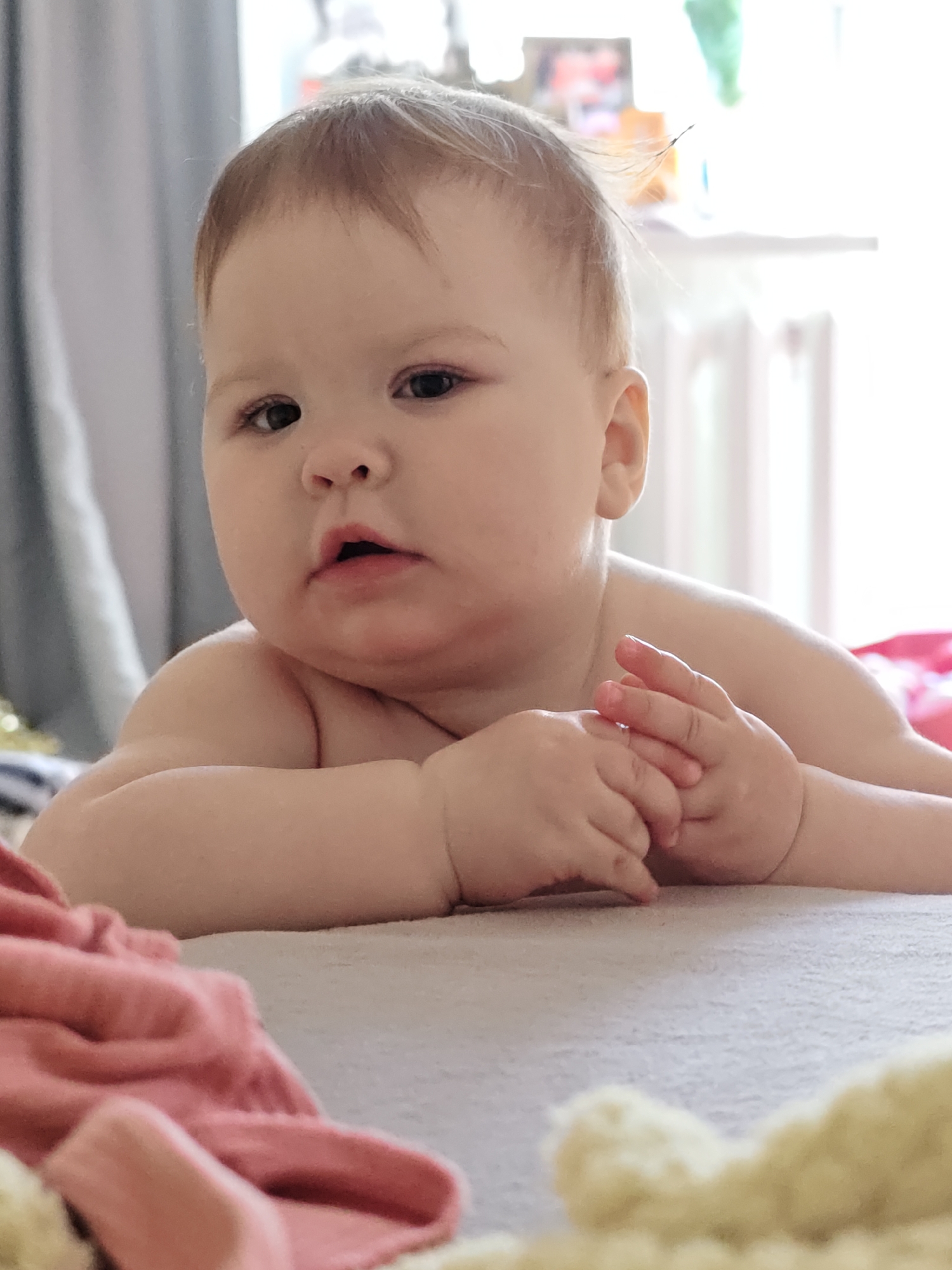 A baby lies on a light-colored surface, possibly a bed, with a pink garment partially visible in the foreground and a window with white blinds and a picture frame in the background. The baby has light skin, brown hair, and is gazing slightly to the left. The baby is holding his/her hands up, with fingers slightly curled. The location is Chaikovsky, Russia.