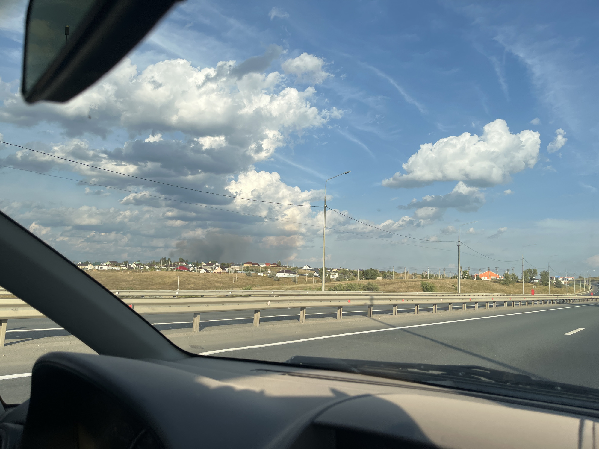 The image depicts a view from inside a vehicle driving on a highway. The road curves to the right and is bordered by a metal guardrail. In the distance, a rural settlement with numerous houses is visible on a gently sloping hill. A plume of smoke rises from the settlement, indicating a potential fire or controlled burn. The sky is a bright blue with scattered white clouds. Power lines run along the horizon. The foreground shows the interior of the car, including the dashboard and a portion of the side mirror. The scene appears to be in or around Dubki, Russia.