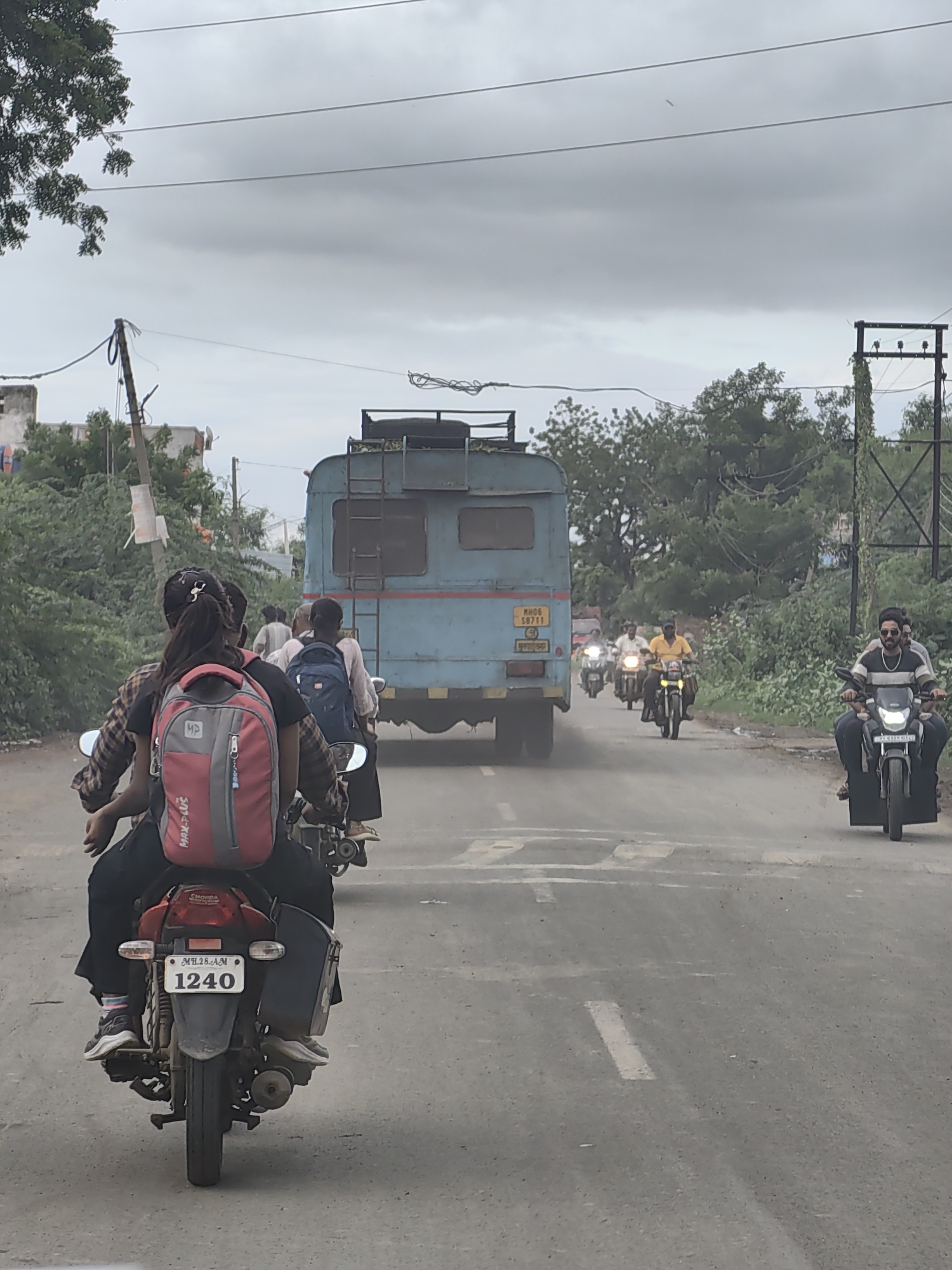 The image shows a street scene in Malkapur, India, with several vehicles on a paved road under a cloudy sky. In the foreground, two people are riding a motorcycle, with the passenger wearing a red and gray backpack. The motorcycle has a license plate reading "MH.28.AM 1240". Behind them, a light blue truck is driving away. Further down the road, multiple other motorcycles with riders are visible. The environment includes trees and vegetation on the sides of the road, and utility poles with wires overhead. The overall impression is of traffic on a day with overcast weather.