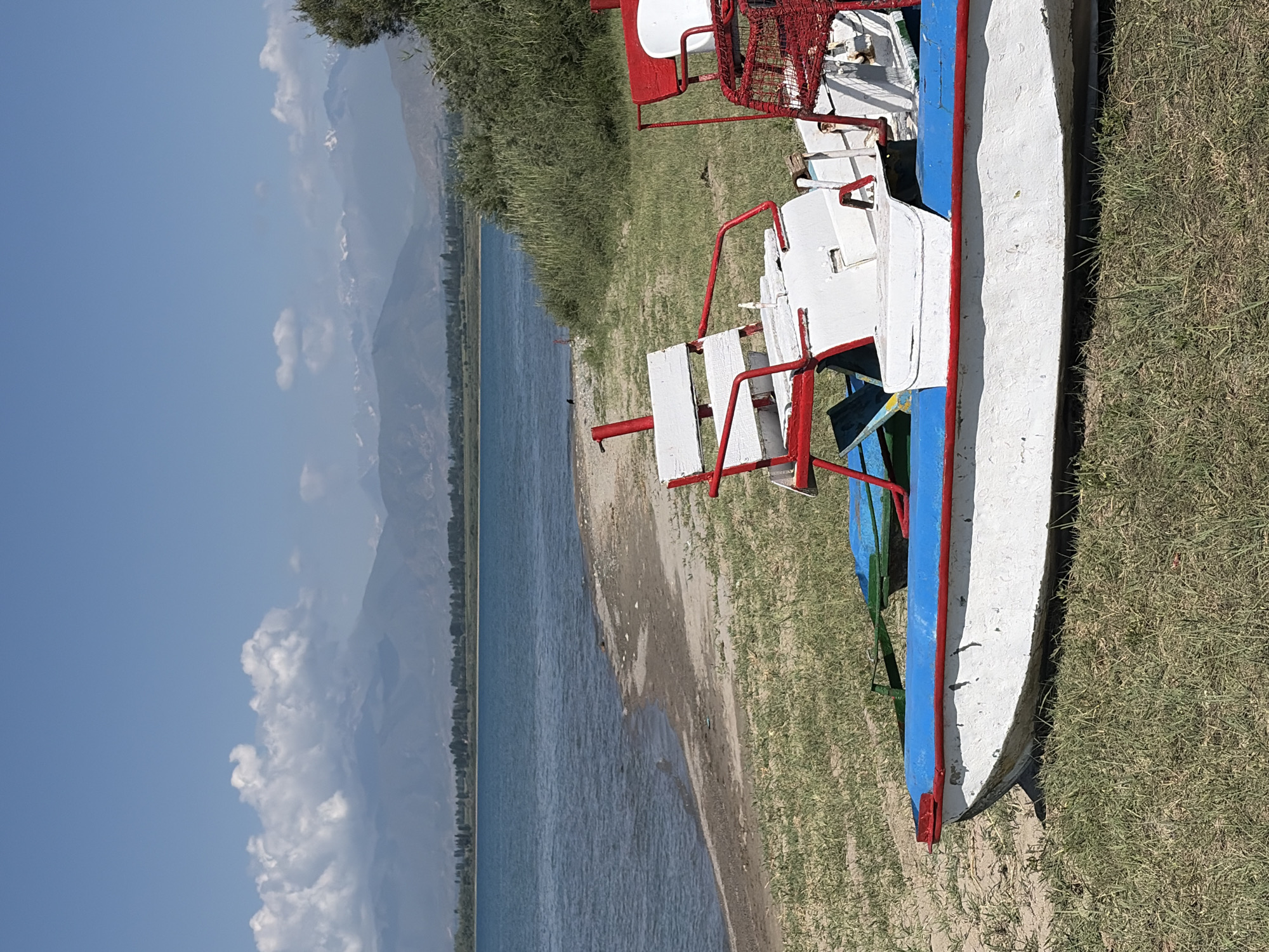 This is a medium shot of a body of water with a beach and some boats in the foreground. The water is a deep blue and calm, with a slight ripple. The beach is sandy and covered with sparse green grass. In the foreground, there are two white and red pedal boats. The boats are partially submerged in the sand and grass, and appear to be weathered. The sky is a clear blue with some white clouds in the distance. In the background, there are mountains. The overall scene is serene and picturesque.