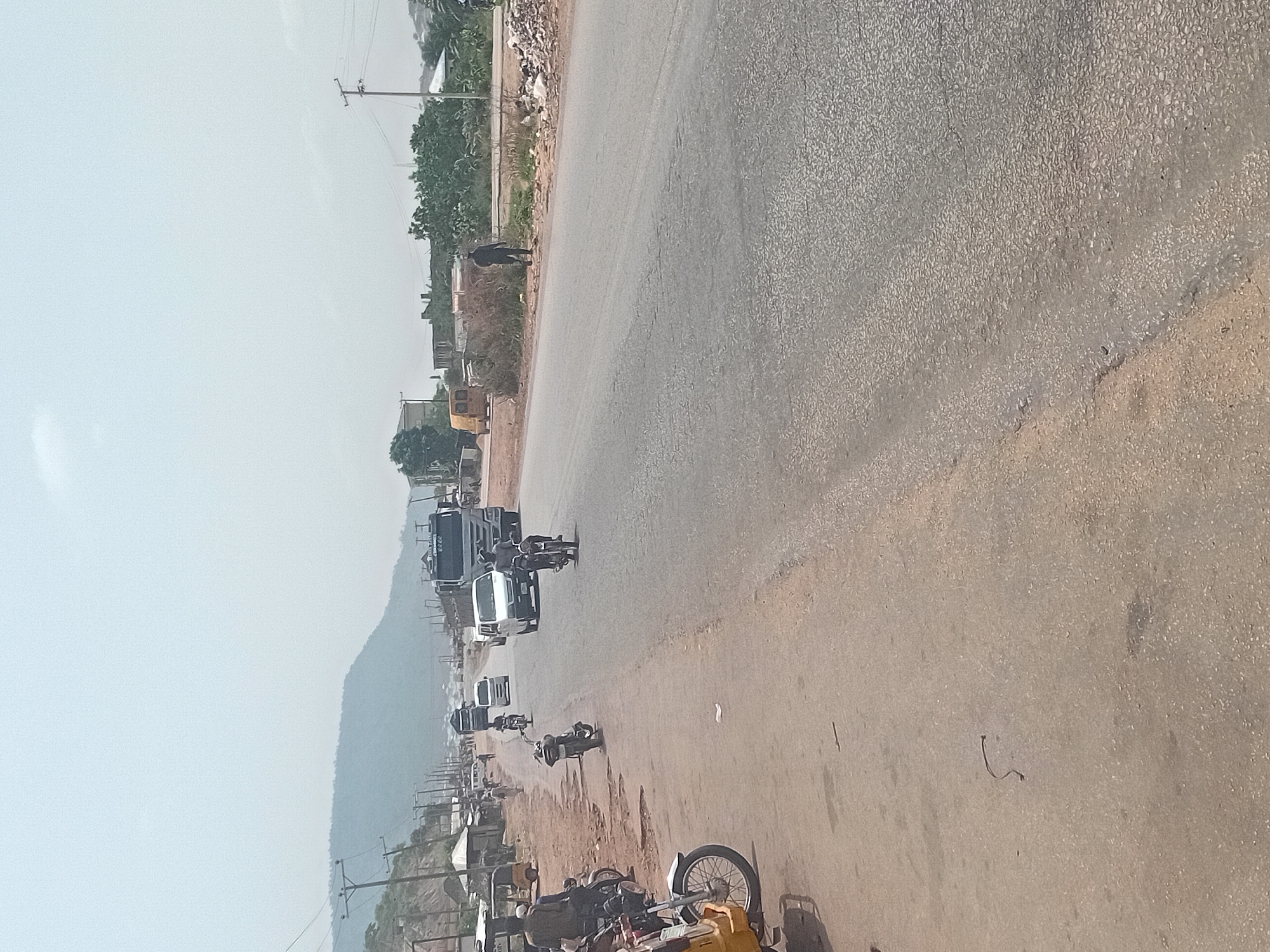 This image depicts a road scene in Lokoja, Nigeria. The road is paved and stretches into the distance with vehicles including a semi-truck and other automobiles traveling on it. On the left side of the road is a shoulder or unpaved area. A mountain range can be seen in the distance. In the foreground, there are motorcycles parked along the road. Power poles line the street. Debris and vegetation are seen along the side of the road. The sky is overcast.