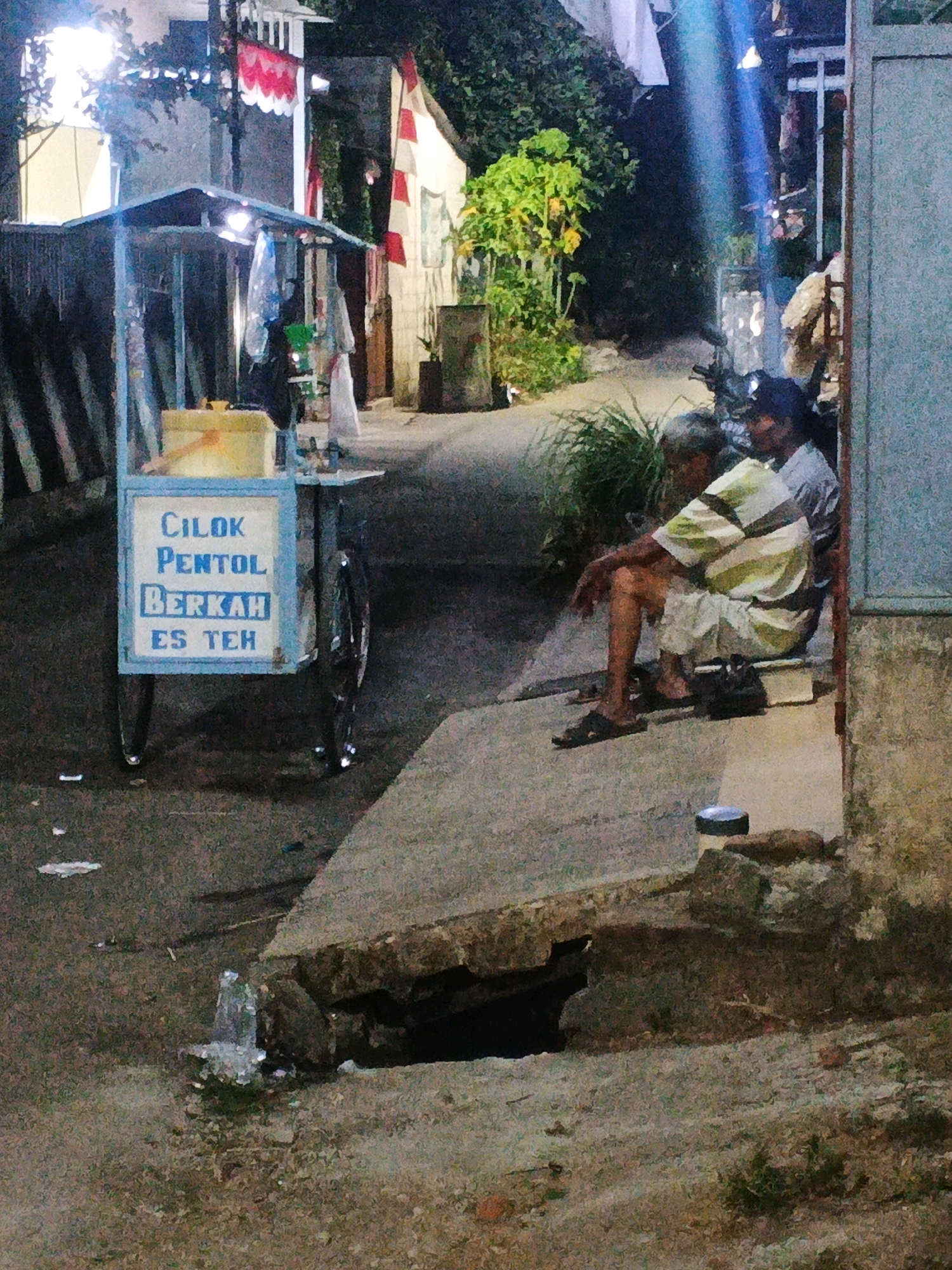The image depicts an outdoor scene at night, possibly in Kadu, Indonesia. A food cart with the text "CILOK PENTOL BERKAH ES TEH" is prominently displayed. Two men are seated on a ledge to the right of the cart. The man in the foreground is wearing a striped shirt and shorts, while the man behind him is wearing a grey shirt. The environment appears to be a street or alleyway with a concrete walkway and a damaged section of pavement in the foreground, revealing a dark cavity. Trees and vegetation are visible in the background. The scene is illuminated by artificial lights, casting shadows and creating a somewhat grainy appearance.