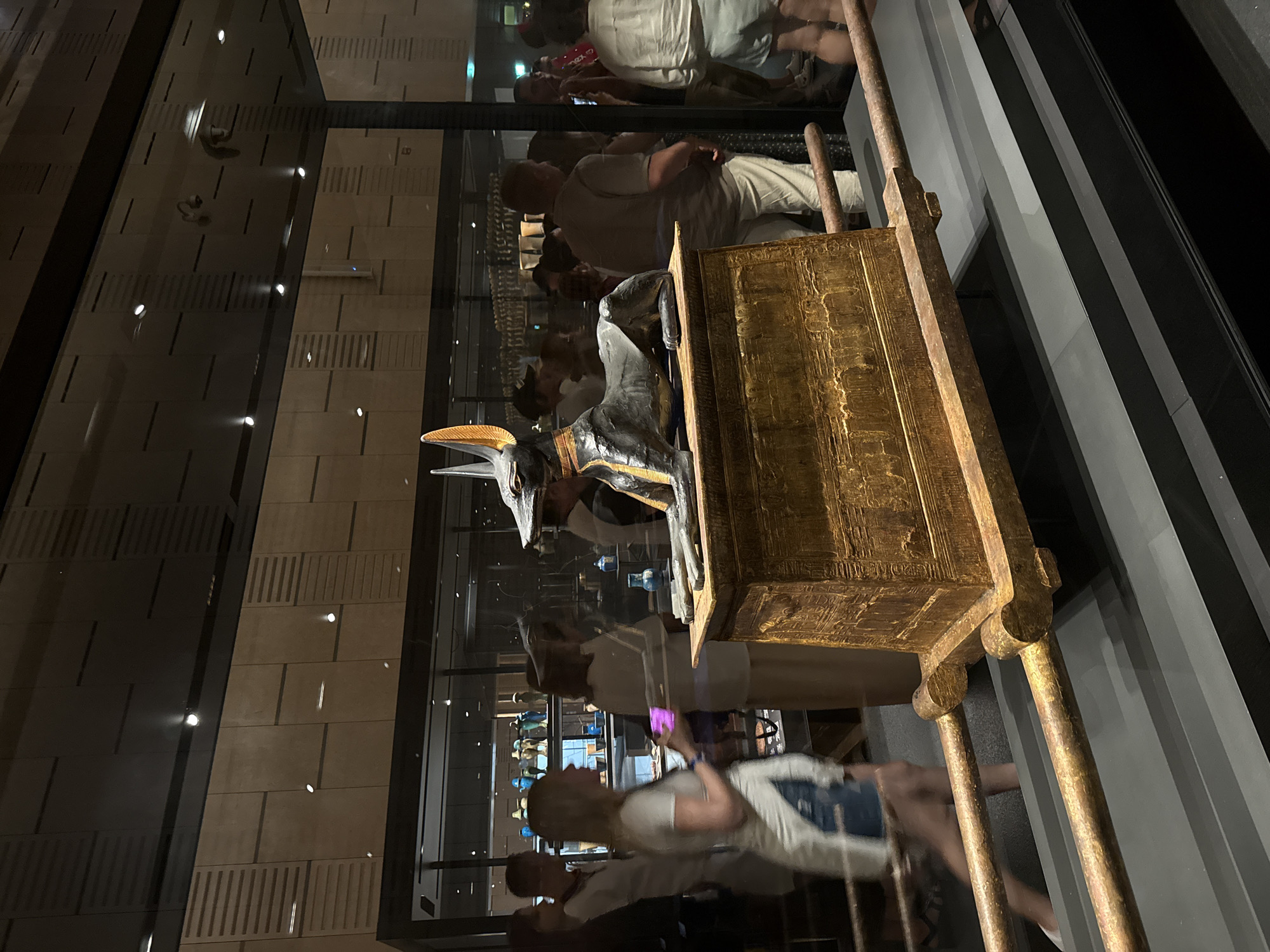 In the city of Kafrat Nassar, Egypt, an ancient Egyptian artifact, specifically a sarcophagus depicting the god Anubis, is displayed within a museum. The sarcophagus, made of wood and gold, rests on a wooden stand with two long, golden poles extending from its sides. The Anubis figure, a jackal-headed deity, is positioned on top of the sarcophagus, its black body contrasting with its golden accents. In the background, several individuals are observed in the museum. One woman in the foreground is wearing a white t-shirt and denim shorts, holding a pink phone. Other museum-goers are visible, some looking at the exhibit, others engaged in conversation. The environment appears to be a dimly lit museum hall, with display cases and lighting fixtures visible. The exhibit is enclosed behind glass.