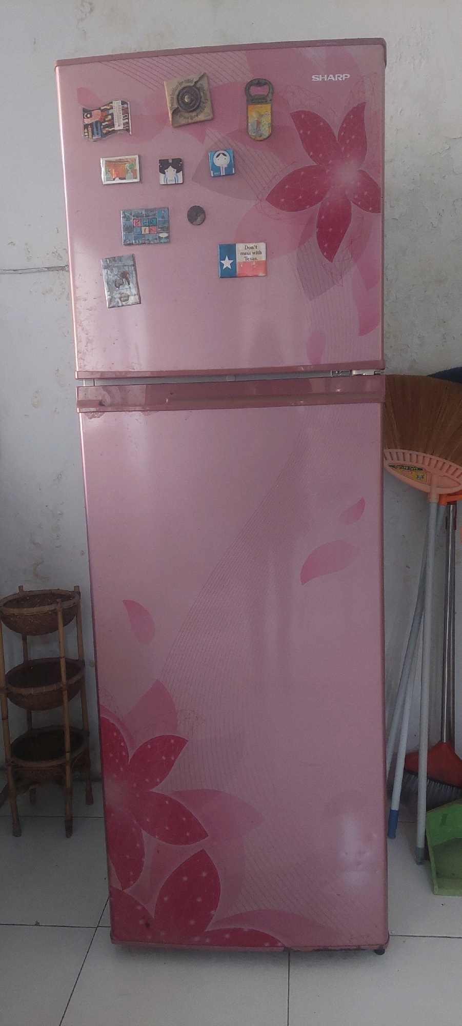 The image shows a pink refrigerator with floral patterns. On the top part of the refrigerator, there are several magnets. One magnet is a flag of Malaysia, another is a picture of a woman and a child, and a bottle opener shaped magnet. There is also a magnet with a star and the text "Don't mess with Texas." The refrigerator is in a room with tiled flooring and a wall that appears to be concrete. To the left of the refrigerator, there is a three-tiered wooden shelf holding woven baskets. To the right of the refrigerator, a broom and dustpan are leaning against the wall. The image does not provide any specific information about the city of Lumajang, Indonesia, or any actions, interactions, or people.