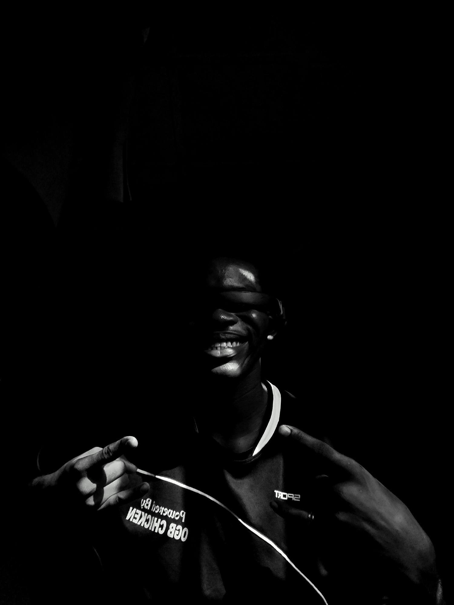 A monochromatic, low-key photograph depicts a smiling man in shadow. Visible are his teeth, the right side of his face from his cheek to his jawline, and a small earring on his right ear. He is wearing a shirt that says "OCB CHICKEN" reversed and "SPORT" partially obscured. He is pointing with both hands towards his chest. A white cord runs from his left hand across his chest and collarbone. The background is predominantly black. It is unknown if the photo was taken in Keffi, Nigeria.