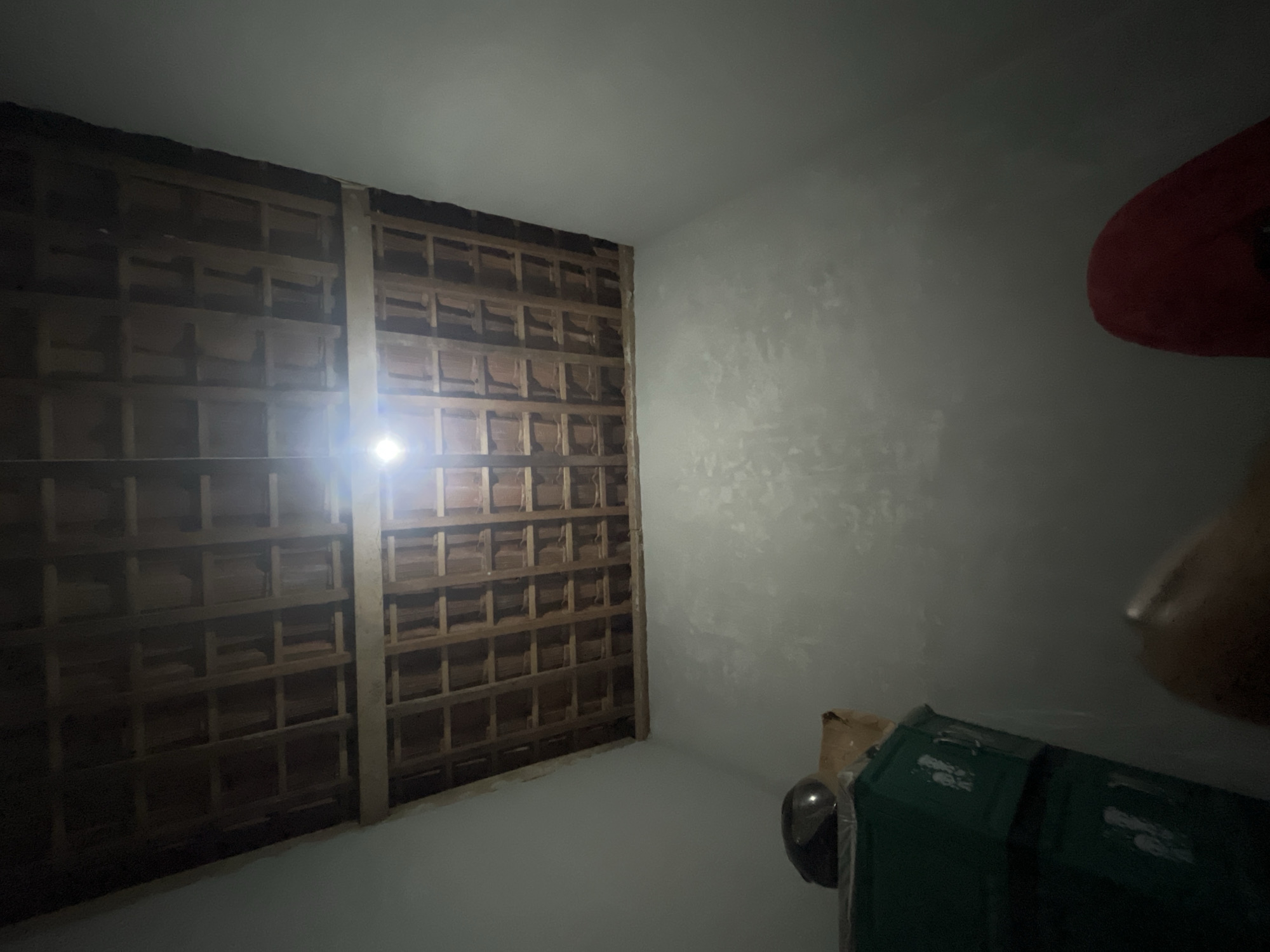 The image shows an interior view of a room with a visible wooden rafter ceiling, possibly under construction or unfinished. A single light bulb hangs from the ceiling, illuminating the space. In the foreground, to the left, are stacked green storage containers. A dark-colored object, possibly a helmet, sits on top of the containers, partially covered by a tan-colored material. To the right and bottom of the frame, a red object, possibly a piece of fabric or clothing, is visible. The walls appear to be plastered or painted in a light, neutral color. The setting is identified as a location in Kebumen, Indonesia. No people are clearly visible in the scene.