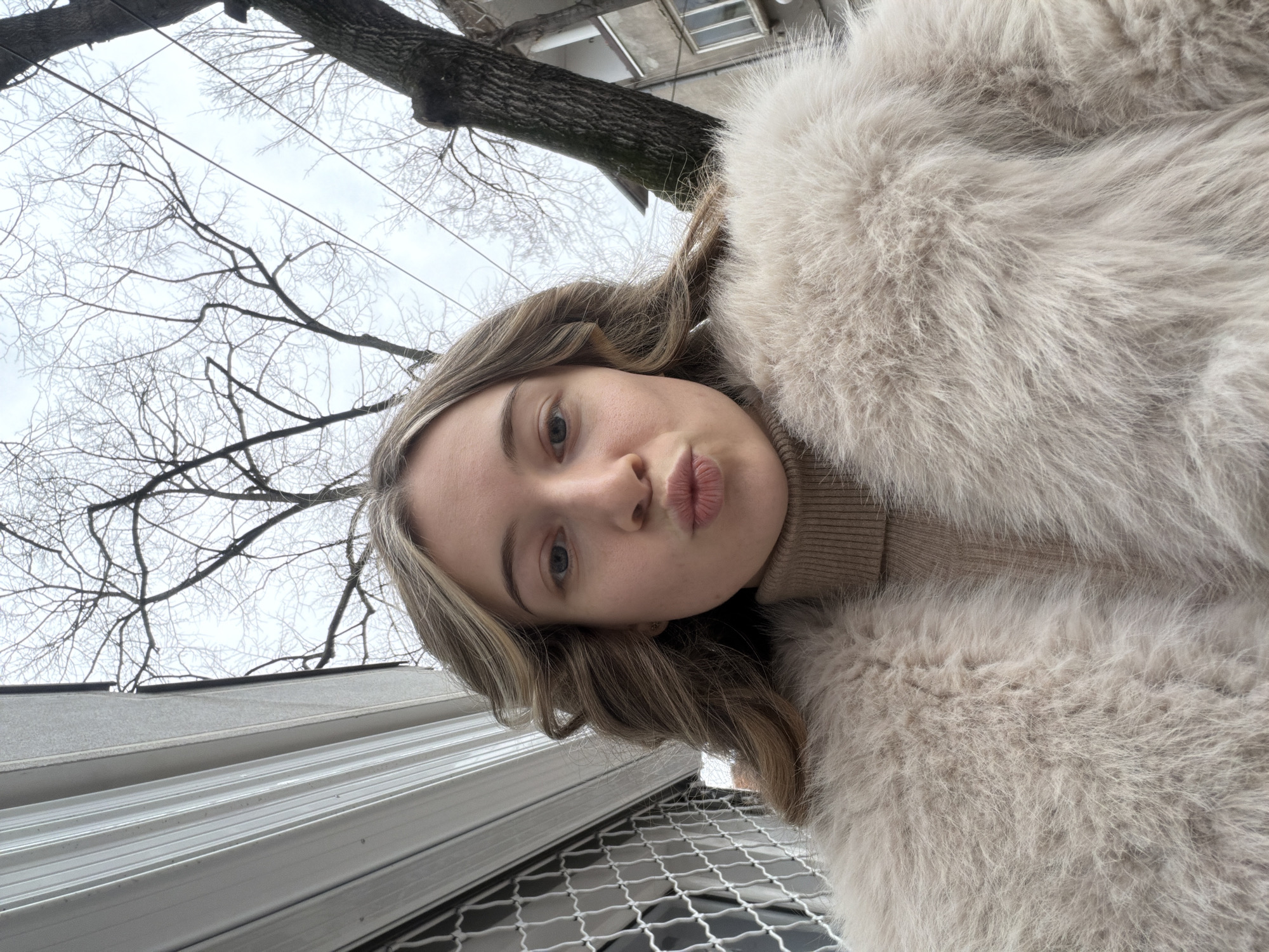 A young woman takes a selfie outdoors. She wears a tan turtleneck sweater and a large, light-colored faux fur coat. Her lips are pursed in a pout. Above and to the left are leafless tree branches against a gray, overcast sky. In the lower left, the side of a white building is visible, along with a dark metal grate. The location appears to be an urban area, but specific details connecting it to Plovdiv, Bulgaria, are not discernible from the image alone.