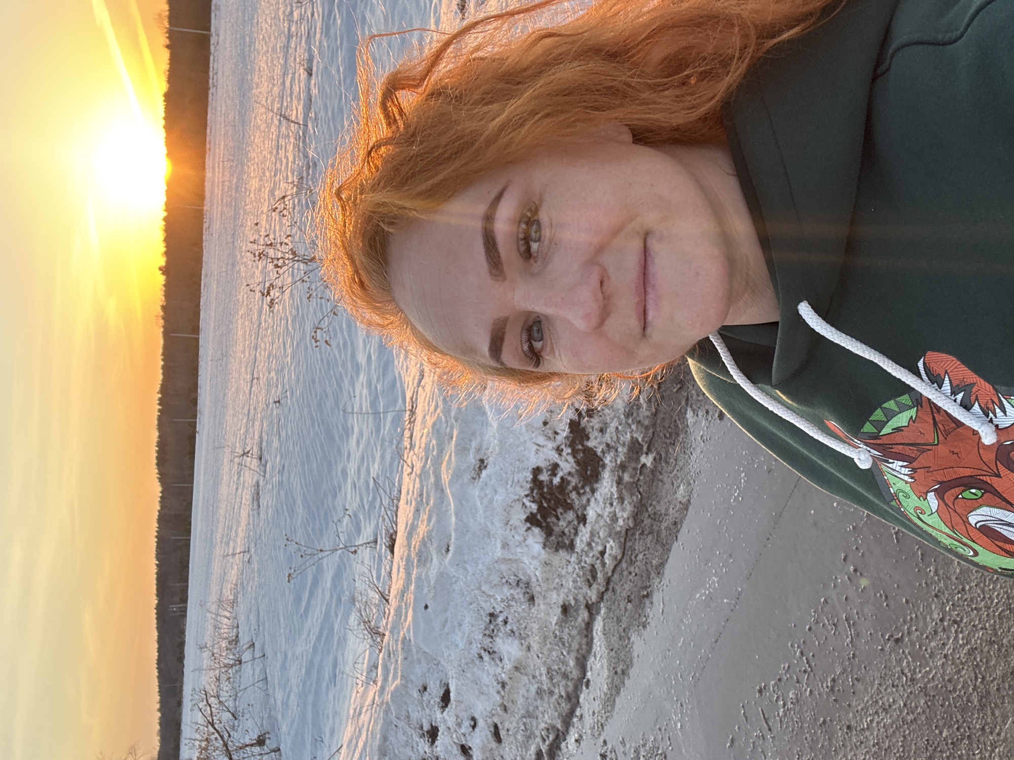 A woman with red hair is taking a selfie at sunset, likely in or near Chaikovsky, Russia, given the location request. She is wearing a green hoodie with a distinctive graphic featuring a fox-like animal. The background consists of a landscape that appears to be covered in snow, with a line of trees silhouetted against the setting sun. The terrain appears rough and uneven in the foreground, suggestive of partially melted snow or ice. The sunset creates a bright, warm light.