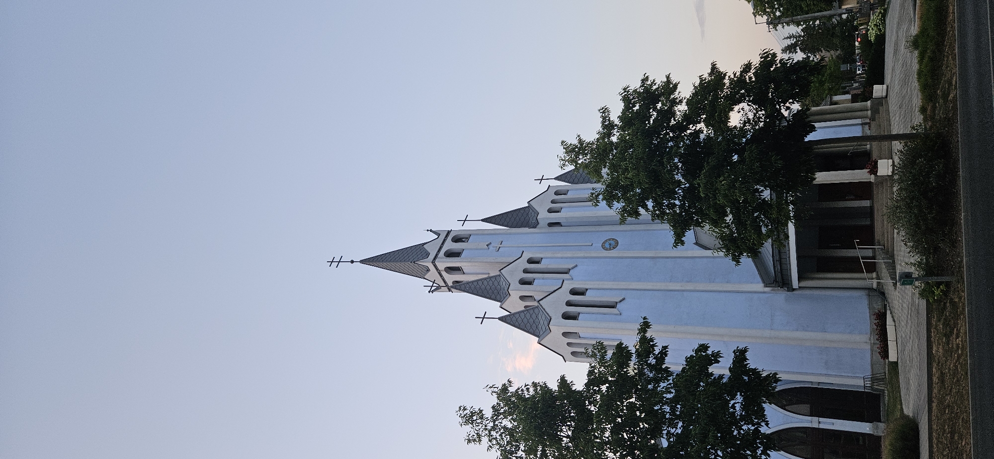 The image shows a blue church with white trim and dark gray spires topped with crosses. The church has a clock on its facade and is surrounded by trees. In the foreground, there is a paved walkway leading to the entrance of the church, with bushes and grass bordering the walkway. In the background, there are more trees and buildings, and a power line. The sky is a clear, pale blue. This is the Catholic Church of the Sacred Heart of Jesus in Hévíz, Hungary.