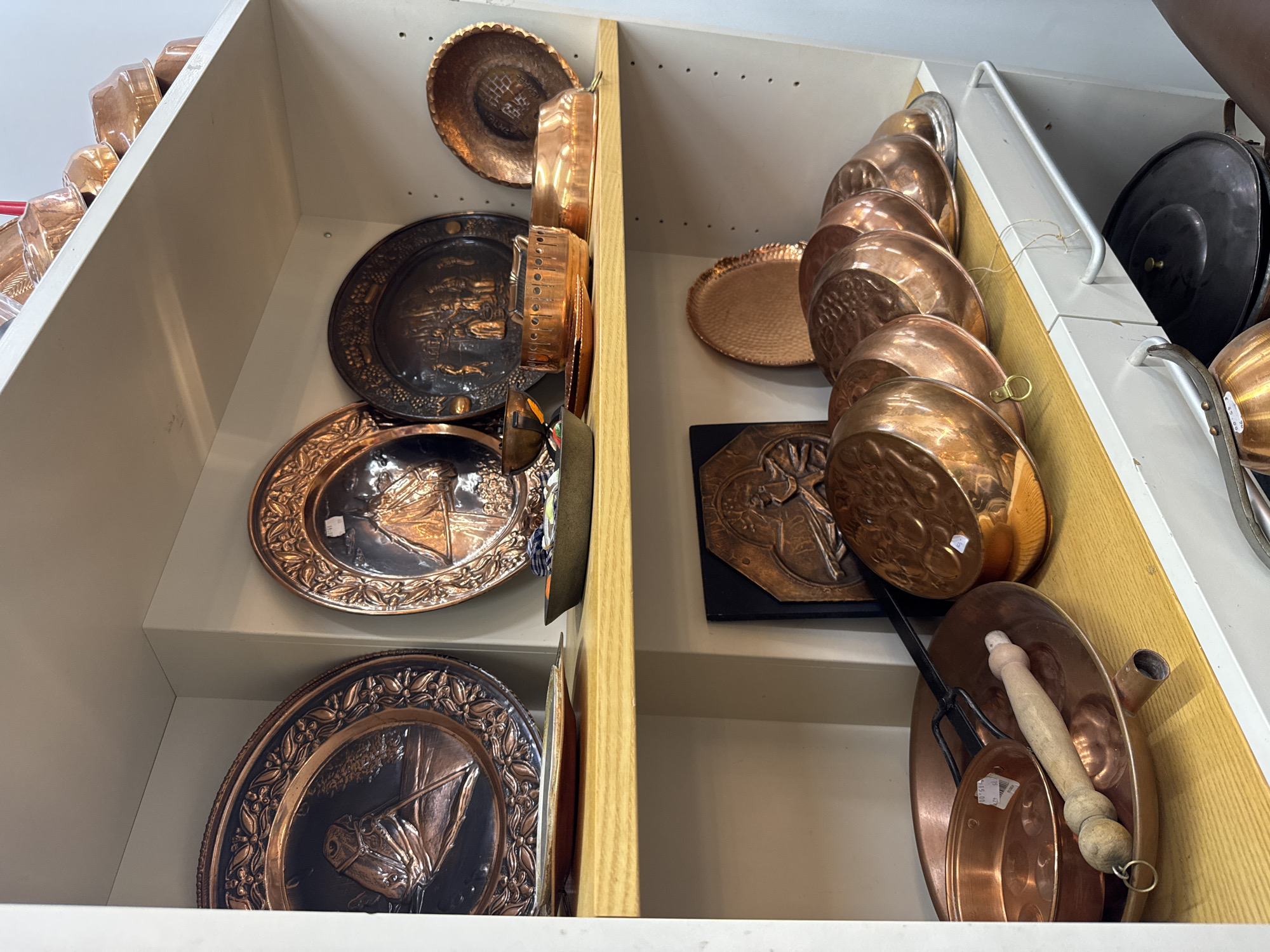 The image shows two shelves inside a cabinet, likely located in Riga, Latvia, displaying a collection of copper and metal items.
The left shelf contains decorative copper plates with embossed designs, stacked vertically. The right shelf displays a variety of copper cookware including pots and pans, also stacked. A square, dark-colored plaque with a copper relief design is visible in the middle of the right shelf. The cabinet is white with a light wood-toned divider. The objects appear to be antiques or vintage items, possibly for sale in a shop.