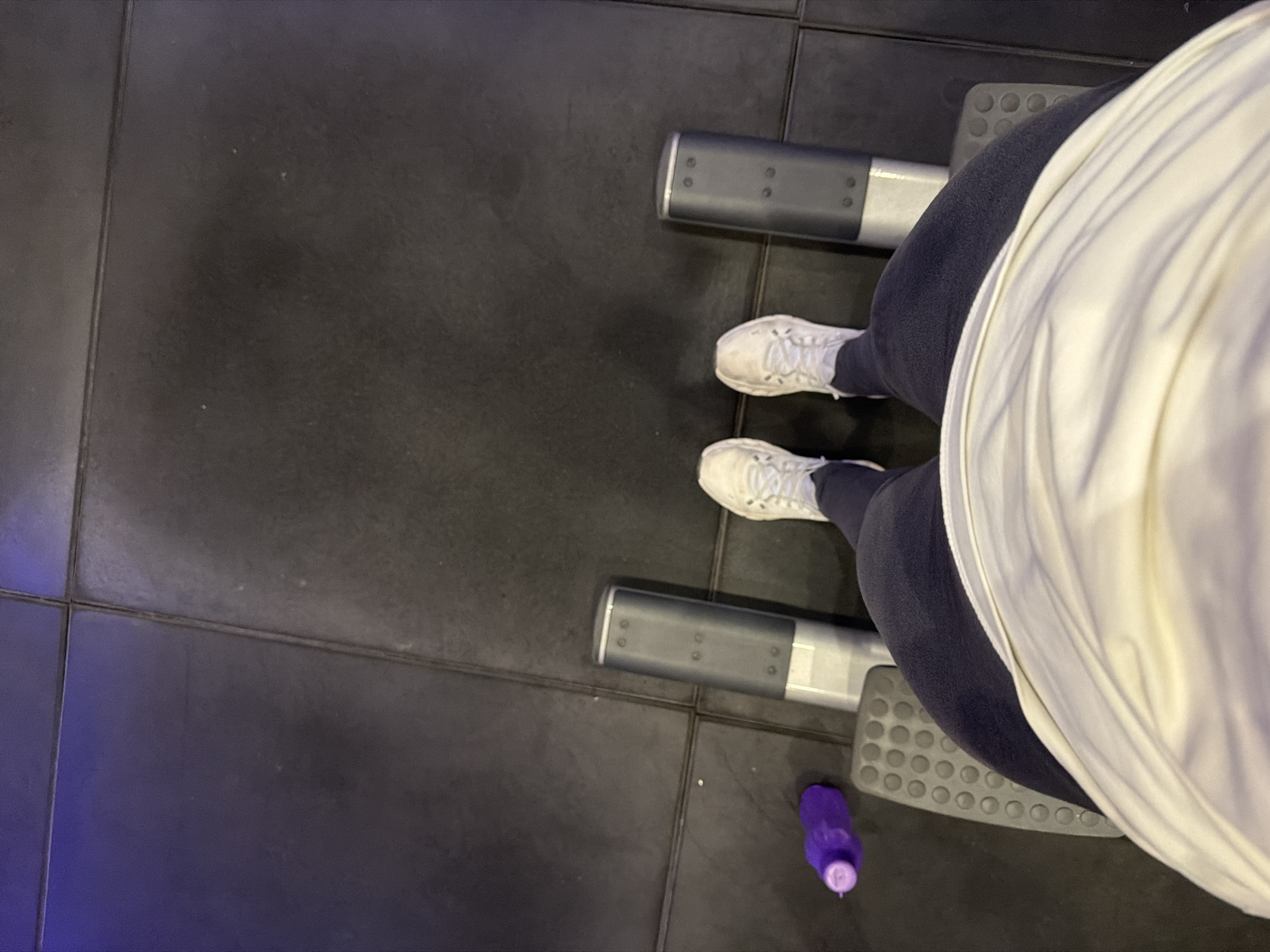 A high-angle shot captures a person on a leg press machine in a gym setting. The person is wearing white sneakers, dark leggings, and a cream-colored shirt or jacket. The leg press machine features gray foot plates and corresponding handles. A small, purple bottle is visible on the floor near the machine. The floor consists of dark gray rectangular tiles. There are no indications of the city of Buenos Aires, Argentina.