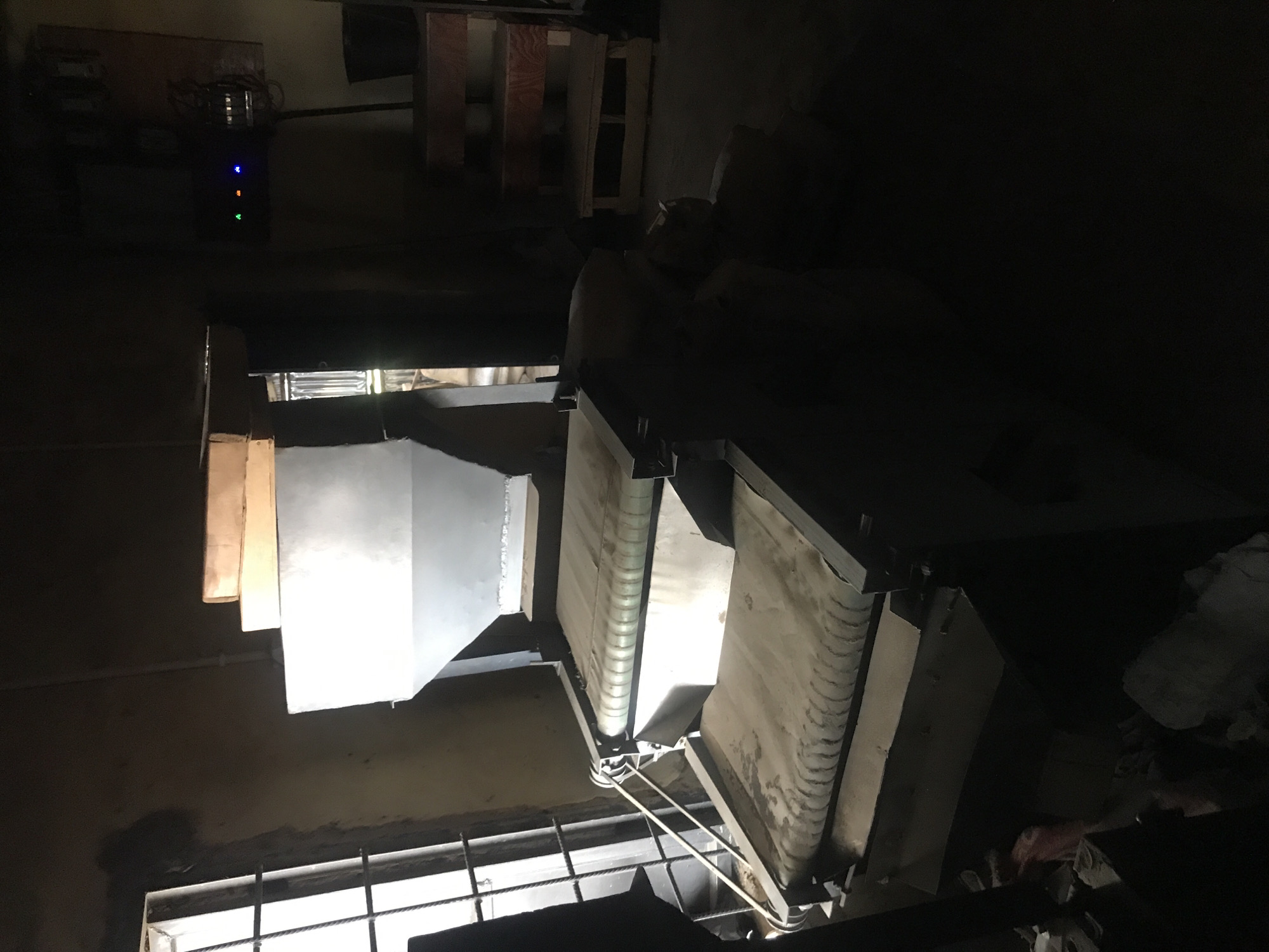 In Kabong, Nigeria, an industrial machine with multiple conveyor belts is visible. The machine is made of metal and appears to be used for processing materials, possibly food or grain, as indicated by the dust and debris on the belts. Above the belts, a large hopper is present, suggesting a feeding mechanism. To the right of the machine, a control panel with illuminated lights is mounted on a wooden board, and a black bucket sits on a wooden shelf. A window with metal bars is situated on the left side of the image, and the overall environment appears to be an indoor industrial or workshop setting with dim lighting.