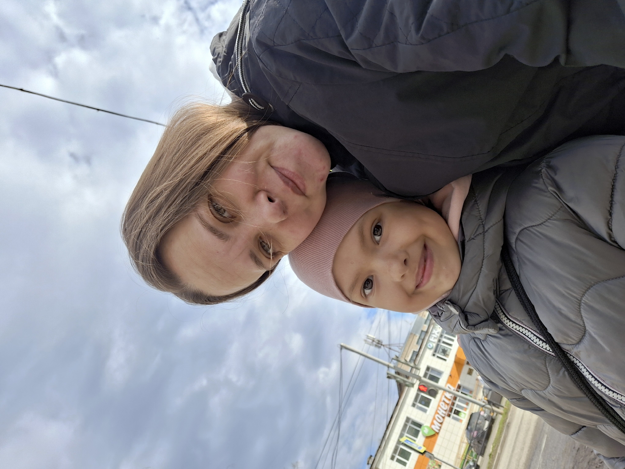 This image is a low-angle selfie, rotated 90 degrees clockwise, featuring an adult woman and a young girl outdoors under a cloudy sky. The adult, with light brown hair and a subtle smile, wears a dark quilted jacket and is positioned in the foreground on the left. Partially behind her and to the right, the young girl, wearing a pink beanie and a grey puffer jacket, also smiles. Both subjects are looking towards the camera, conveying happiness.
The background reveals an urban or suburban setting, consistent with Chaikovsky, Russia. Overhead wires stretch across the overcast sky. Buildings line a street in the mid-ground, including one prominently displaying Cyrillic text: "МОНЕТКА" (Monetka), a well-known Russian supermarket chain, and what appears to be "ФИКС" (Fix) below it. A red traffic light and other street infrastructure are also visible. The scene takes place during daytime with overcast weather.