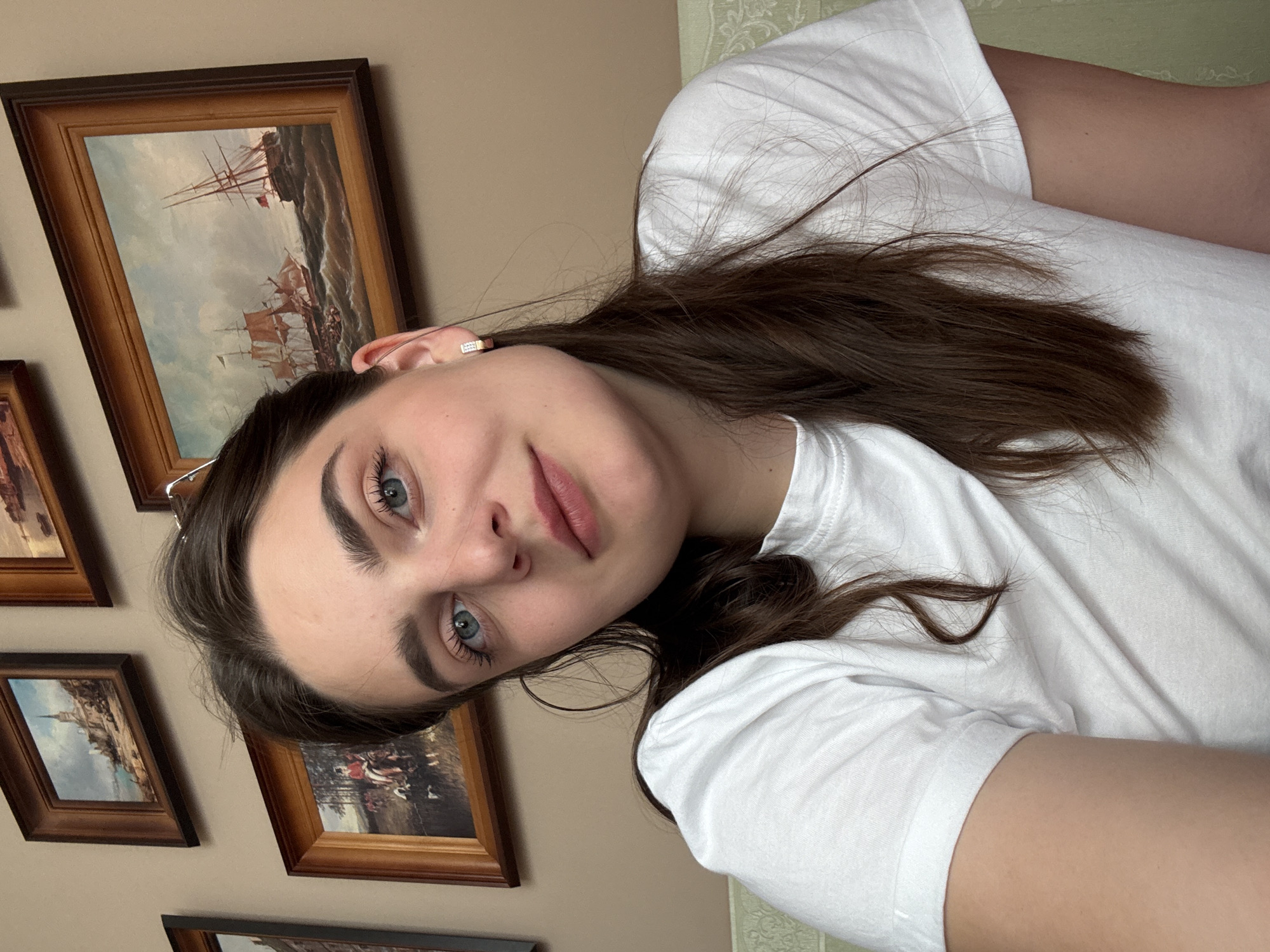 A young woman is posing for a photo indoors, wearing a white T-shirt. She has brown hair and blue eyes. Behind her, on the wall, are multiple framed paintings. The background wall is beige. The specific location in Trnava, Slovakia, cannot be determined from the given image.