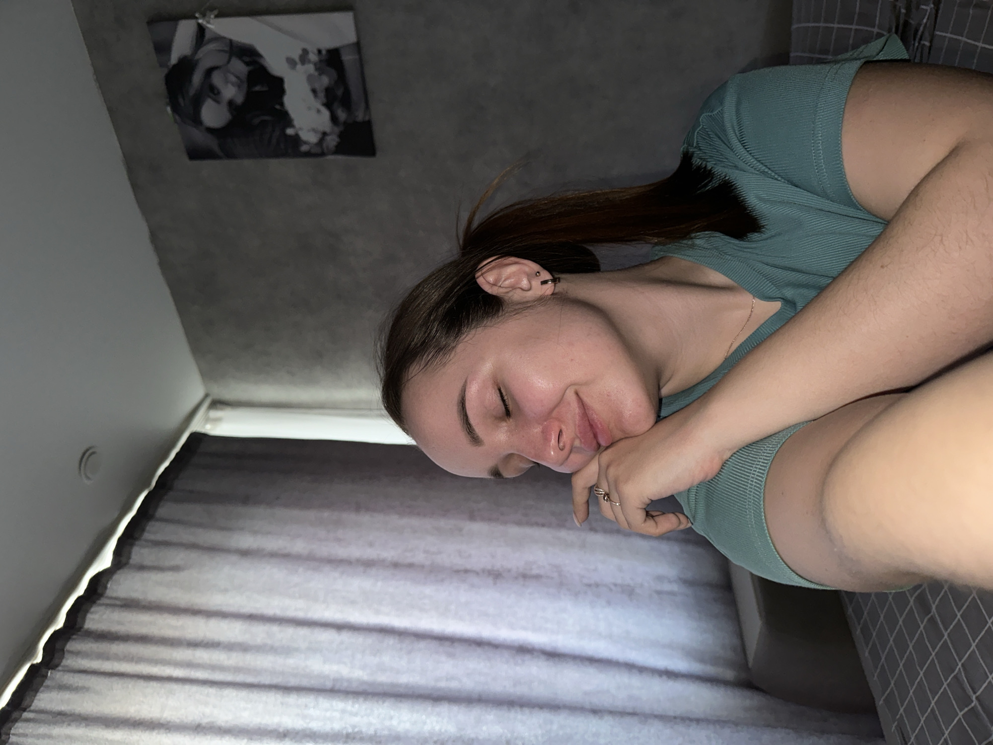 The image shows a young woman with light skin and dark brown hair pulled back into a ponytail. She is wearing a turquoise ribbed tank top and resting her cheek on her hand, which has a ring on the ring finger. The background includes a gray wall with a framed black and white photo hanging on it and light gray curtains. She appears to be lying in bed with gray and white checkered sheets visible on the right side. The location is possibly Chaikovsky, Russia.