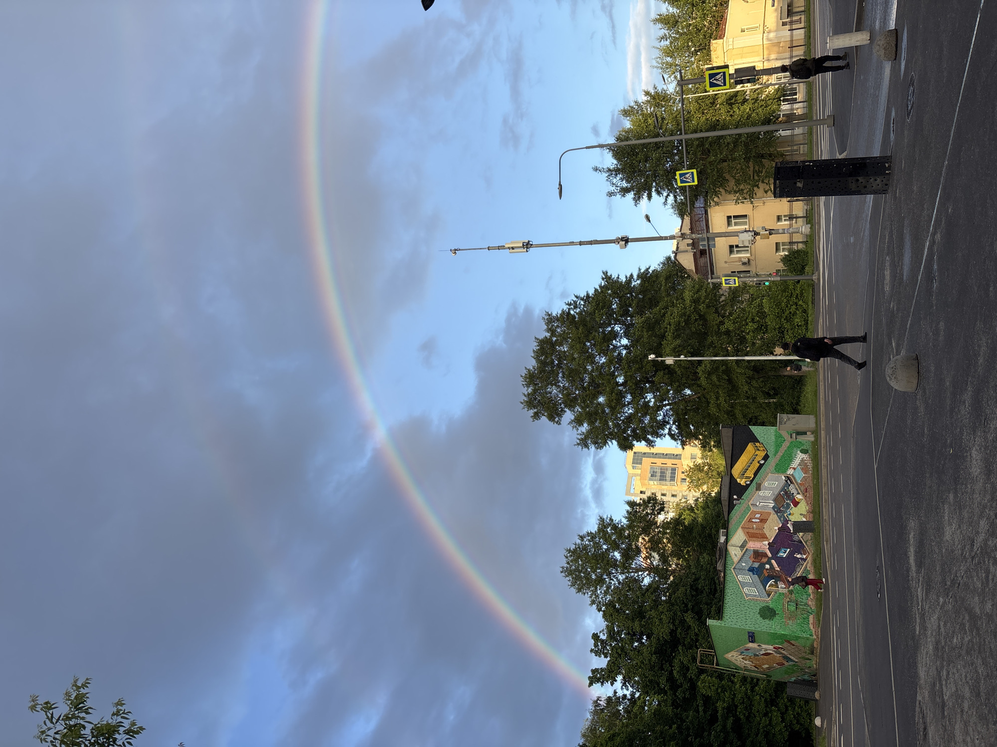 A double rainbow arcs across a partly cloudy sky, with the city of Moscow, Russia visible below. The rainbow extends from the upper left to the lower right. The cityscape includes buildings of varying heights and styles, some with green foliage. A mural depicting colorful houses and a yellow school bus is visible on a building on the right side of the frame. Streetlights and pedestrian crossing signs stand along a street with two people walking on opposite sides. The road is asphalt. A black metal structure with circular perforations is situated on the sidewalk.