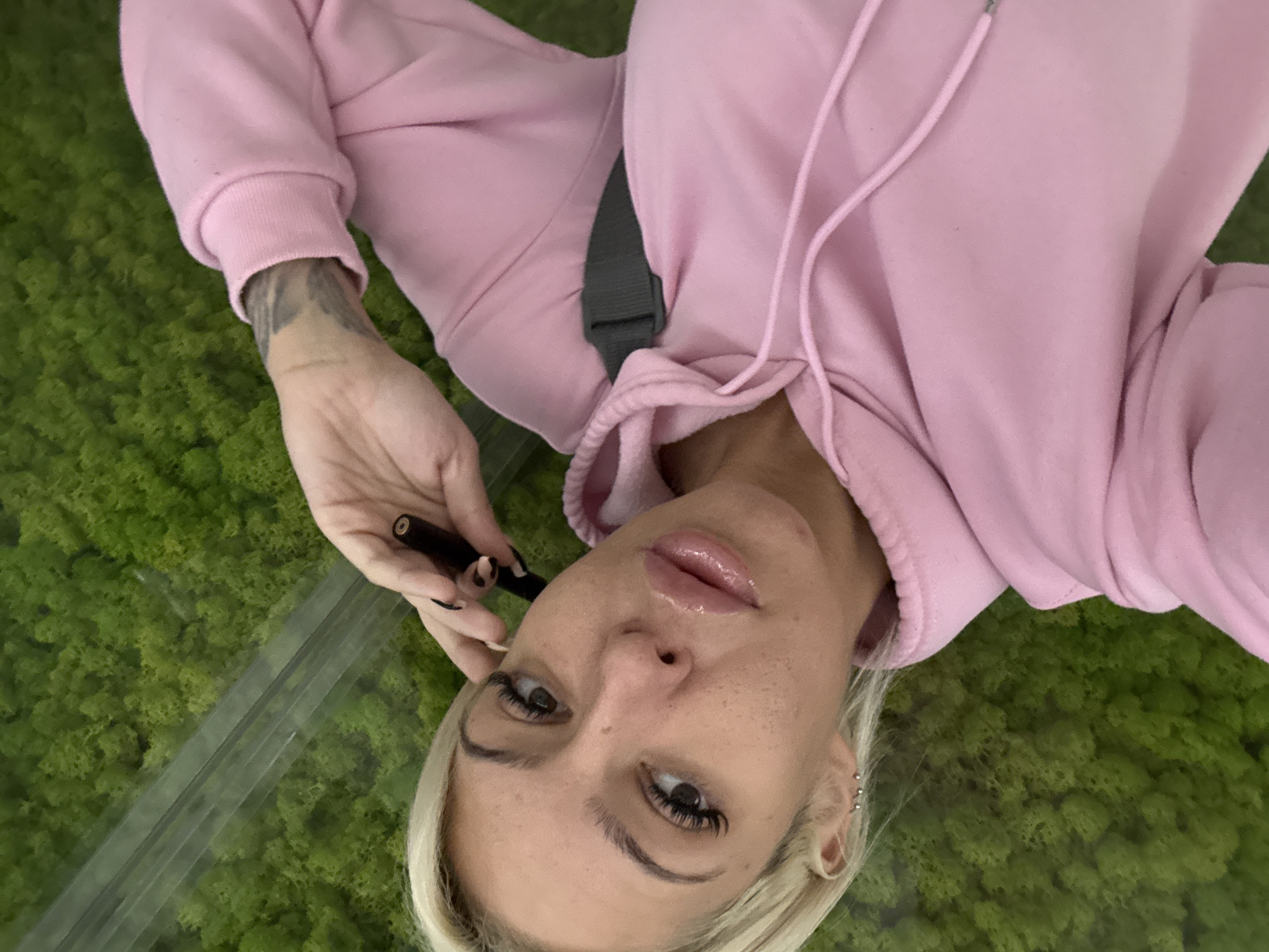 A medium shot captures a woman from a slightly elevated perspective, as if the viewer is looking down upon her. She lies on a vibrant green surface, possibly moss, that extends behind her. A transparent, linear object, perhaps glass, is positioned on the left.
The woman is fair-skinned with blonde hair and noticeable freckles. She is adorned with eyelashes and has multiple piercings in her ear. She is wearing a pink hooded sweatshirt. Her left arm is visible, revealing a tattoo on her forearm. She holds a dark-colored tube, possibly a cosmetic item, in her left hand. A gray strap crosses her body.
The location and city are not discernible from the image.