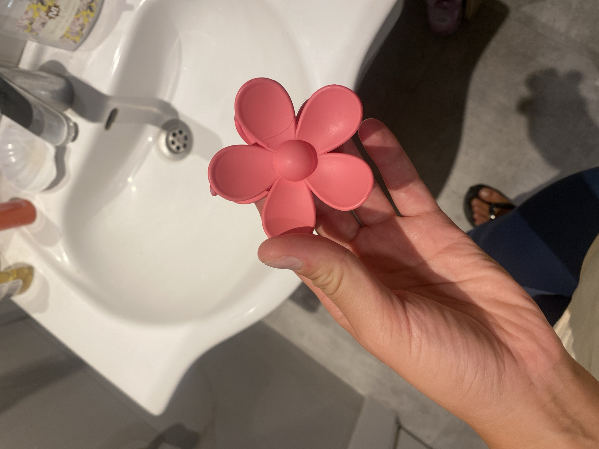 A hand holds a pink, flower-shaped hair claw over a white ceramic sink. The sink has a chrome faucet and drain visible. Various toiletries, including a bottle with a yellow cap and a clear bottle with a floral label, are on the edge of the sink. The floor appears to be a light gray tiled surface. The scene suggests a bathroom or wash area. The location is not provided in the image.