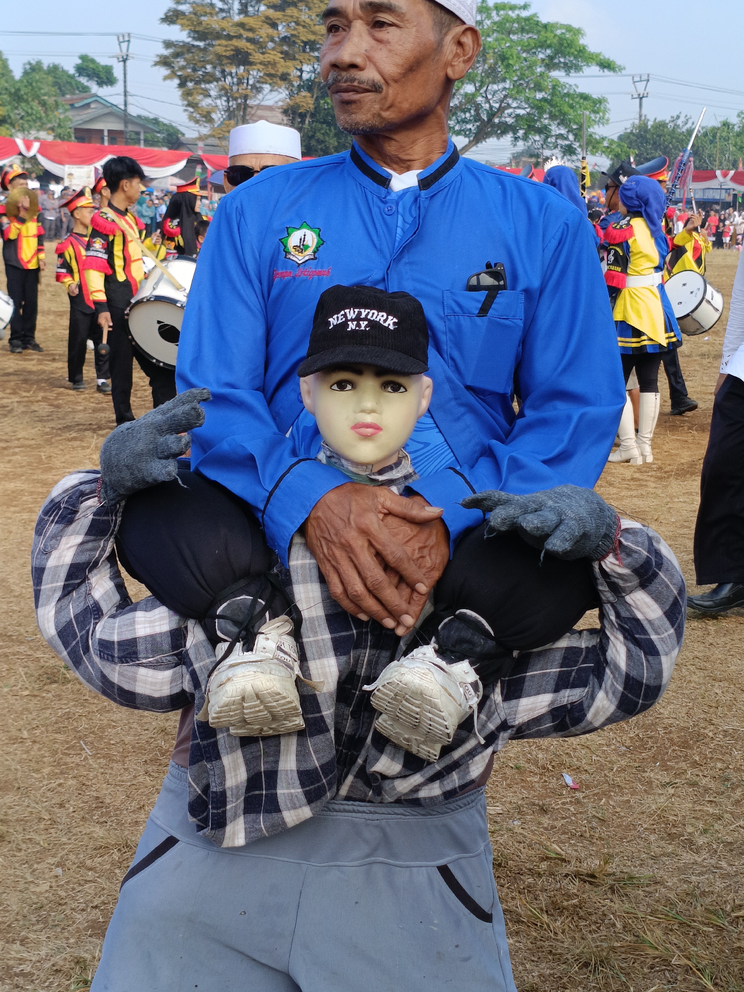 In Cibodas, Indonesia, an elderly man in a blue shirt and white cap is holding a mannequin. The mannequin, wearing a plaid shirt, grey gloves, and sneakers, is seated on the man's lap, with its legs positioned as if sitting on the man's shoulders. The man's hands are around the mannequin's waist. Behind them, a marching band in colorful uniforms is visible, some members holding drums and flags, suggesting an outdoor event or parade. The ground is covered with dry grass and scattered debris.