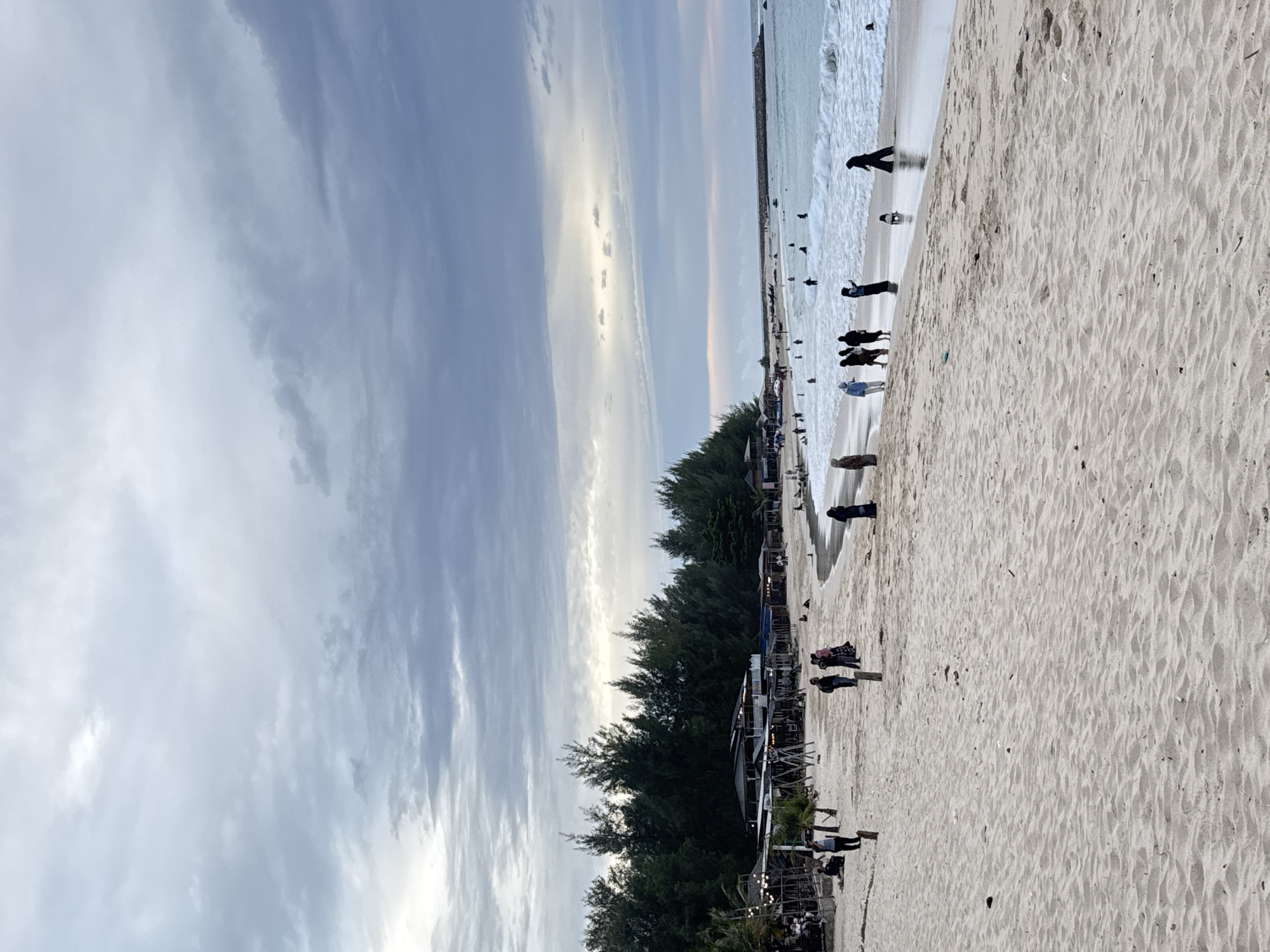 The image depicts a crowded beach scene at dusk in Meunasahbale, Indonesia. A line of trees borders the sandy expanse, behind which a row of small establishments with thatched roofs are visible, likely shops or food stalls. Several people are walking along the beach, some are standing near the water, and a few are in the ocean, wading through the waves. The sky is overcast with dramatic clouds, hinting at the end of the day or impending weather. The waves are moderately sized, creating a white foamy crest as they approach the shore. The sand is light in color and shows signs of being slightly damp near the water's edge.