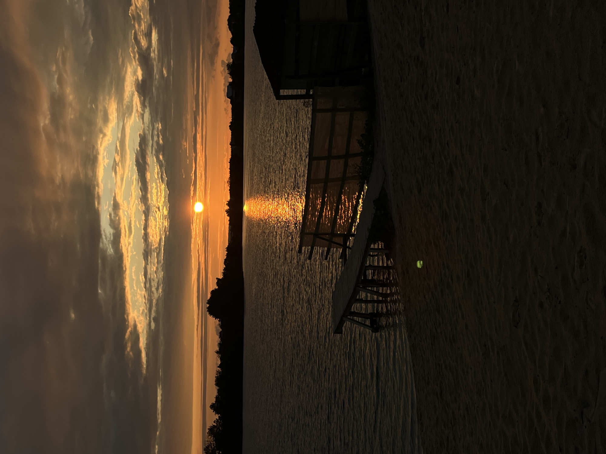 A serene sunset is captured over the water at Bezliudivka, Ukraine. The sun, a brilliant orb of orange, is low on the horizon, casting a warm glow across the sky and reflecting on the rippling surface of the lake. Dark, dramatic clouds streak across the upper part of the frame, their undersides illuminated by the setting sun. Along the distant shore, a silhouetted treeline stretches across the landscape, with a few faint structures visible. In the foreground, a sandy beach leads down to the water's edge. A rustic wooden pier juts out into the lake, its structure dark against the illuminated water. To the right of the pier, a small, simple wooden structure is partially visible, set back from the water. A single yellow tennis ball rests on the sand near the water. The scene is peaceful and tranquil, evoking the quiet beauty of a summer evening.