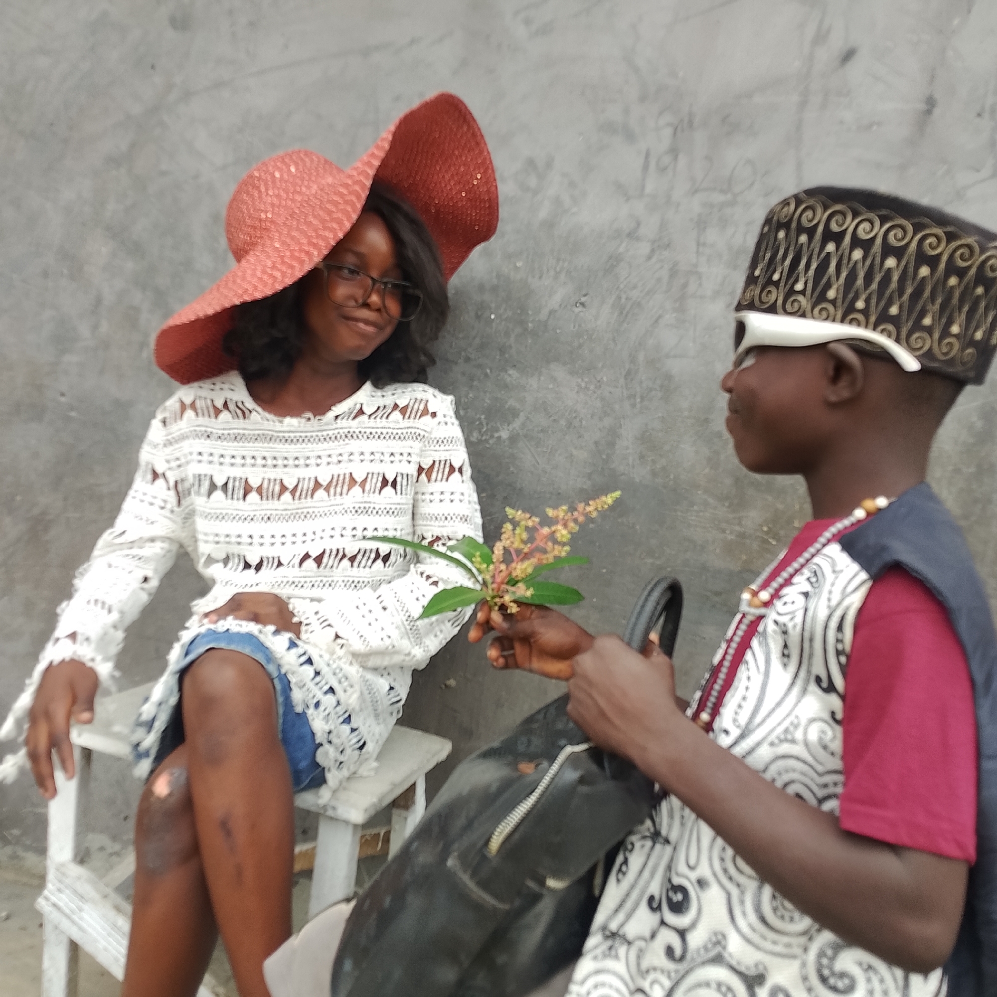 In Ado, Nigeria, two young people are posed against a concrete wall. The individual on the left, wearing a large, pink, wide-brimmed hat and glasses, is dressed in a white, intricately crocheted garment over denim shorts. They are seated on a white wooden bench. The other individual, on the right, is wearing a black cap with ornate gold patterns, white-rimmed glasses, and a beaded necklace. They are also wearing a white and black patterned garment with a maroon undershirt and a dark blue over-garment. This individual is holding a branch with small flowers and leaves, extending it towards the person on the left. A black bag rests on the lap of the person on the right. The background is a textured, gray concrete wall.